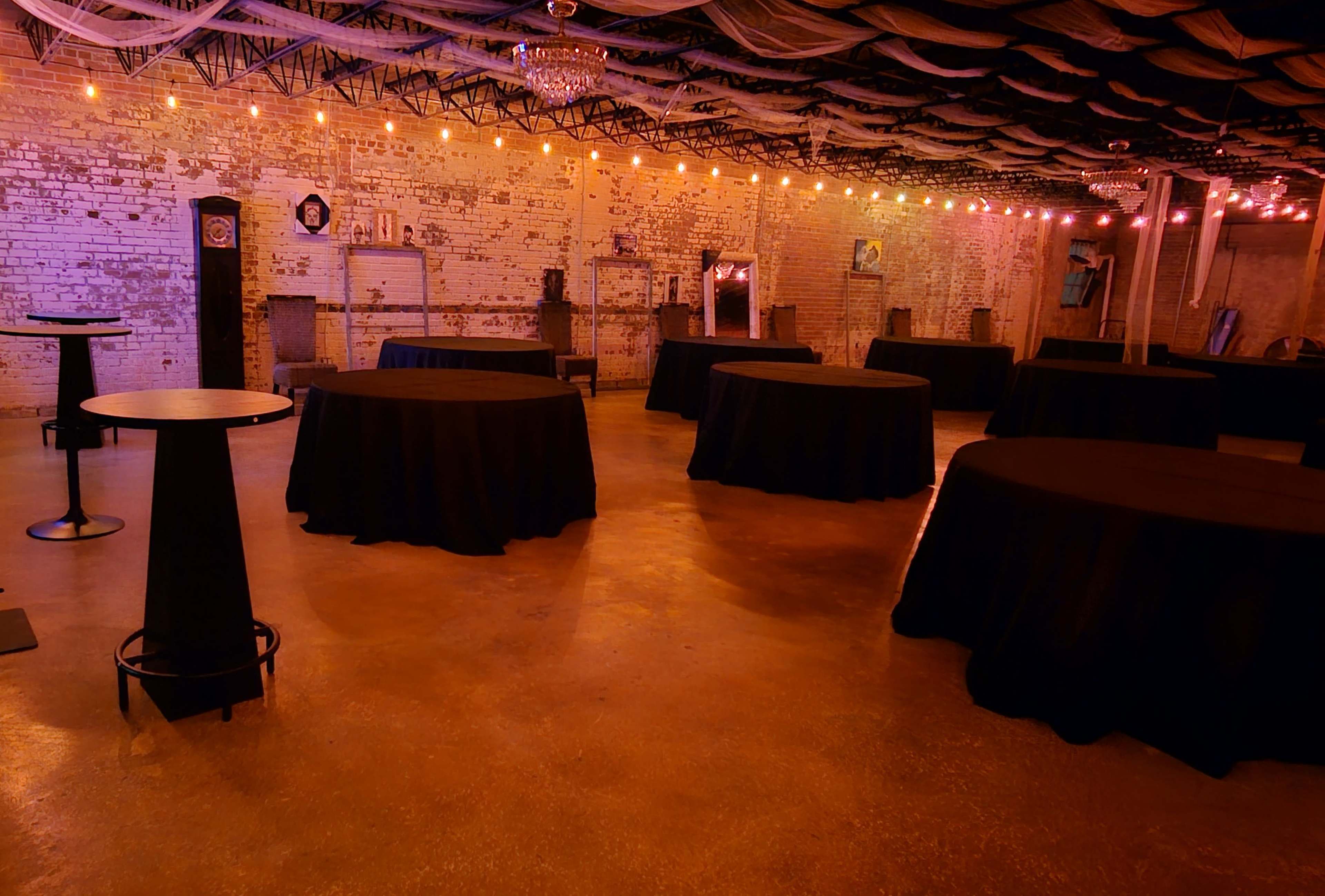 The image shows an event space with several round tables covered in black tablecloths, surrounded by exposed brick walls and warm string lights.