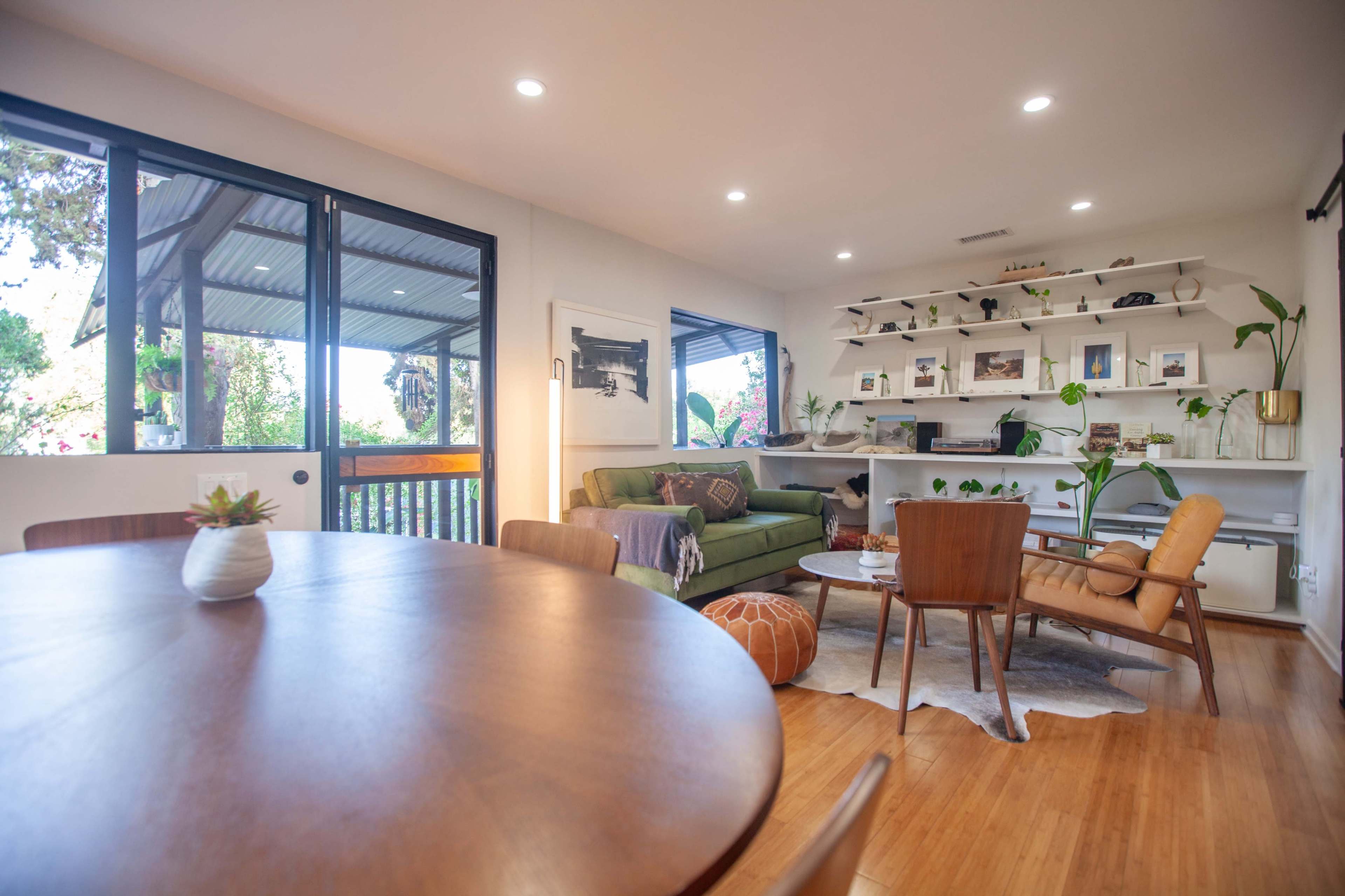 A bright living area features a green couch, a round dining table, and a shelf filled with decorative items and plants.