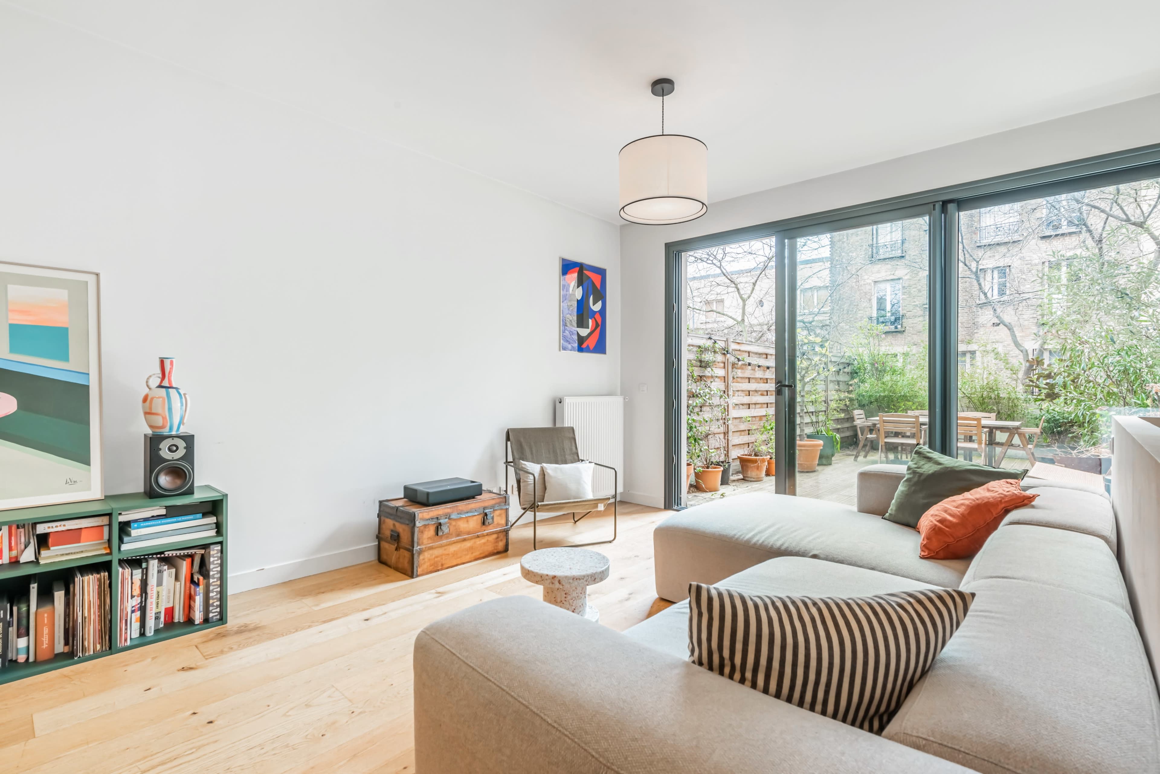 A modern living room features a light-colored sofa, a wooden floor, and large glass doors opening to a garden area.
