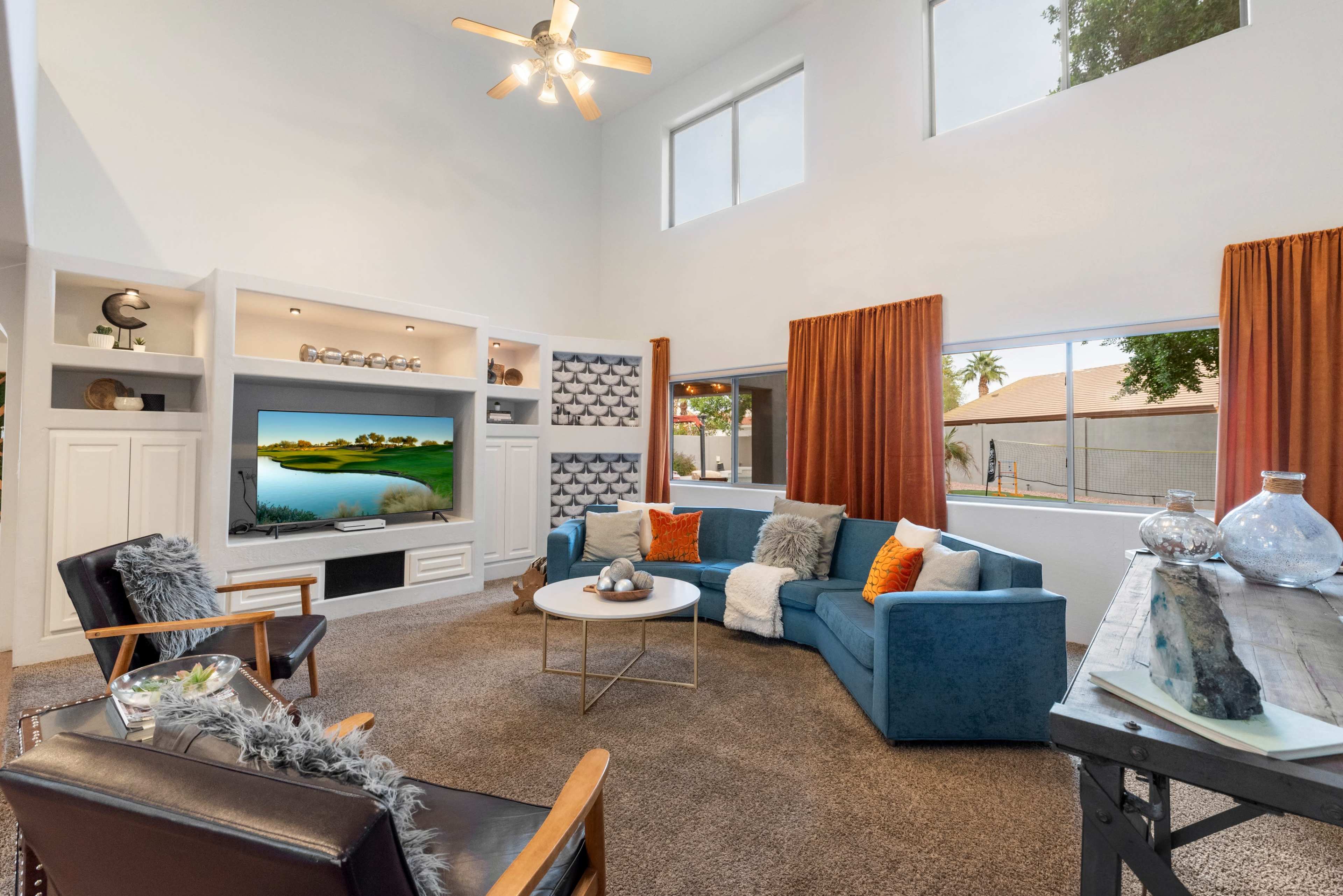 A modern living room features a blue sectional sofa, two wooden chairs, a round coffee table, and a mounted TV, with large windows allowing natural light to illuminate the space.