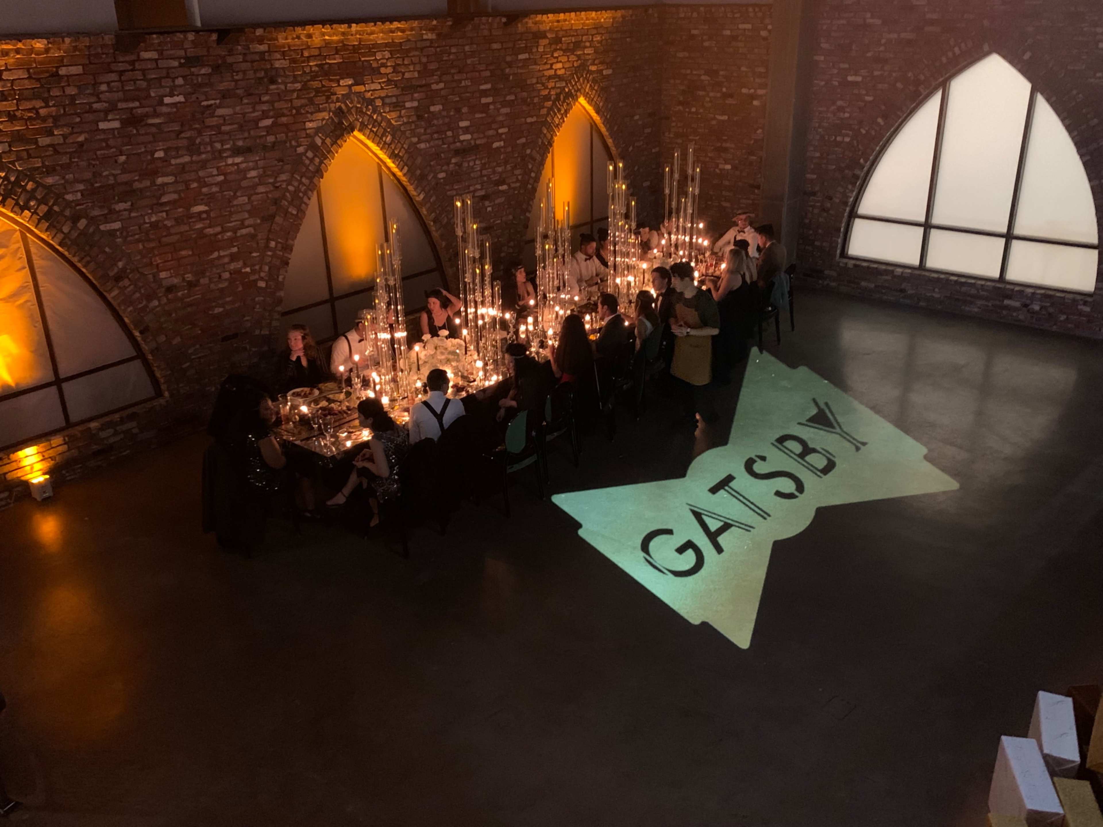 A long dinner table is set with candles in a dimly lit room featuring brick walls and arched windows, with the word "GATSBY" projected on the floor.