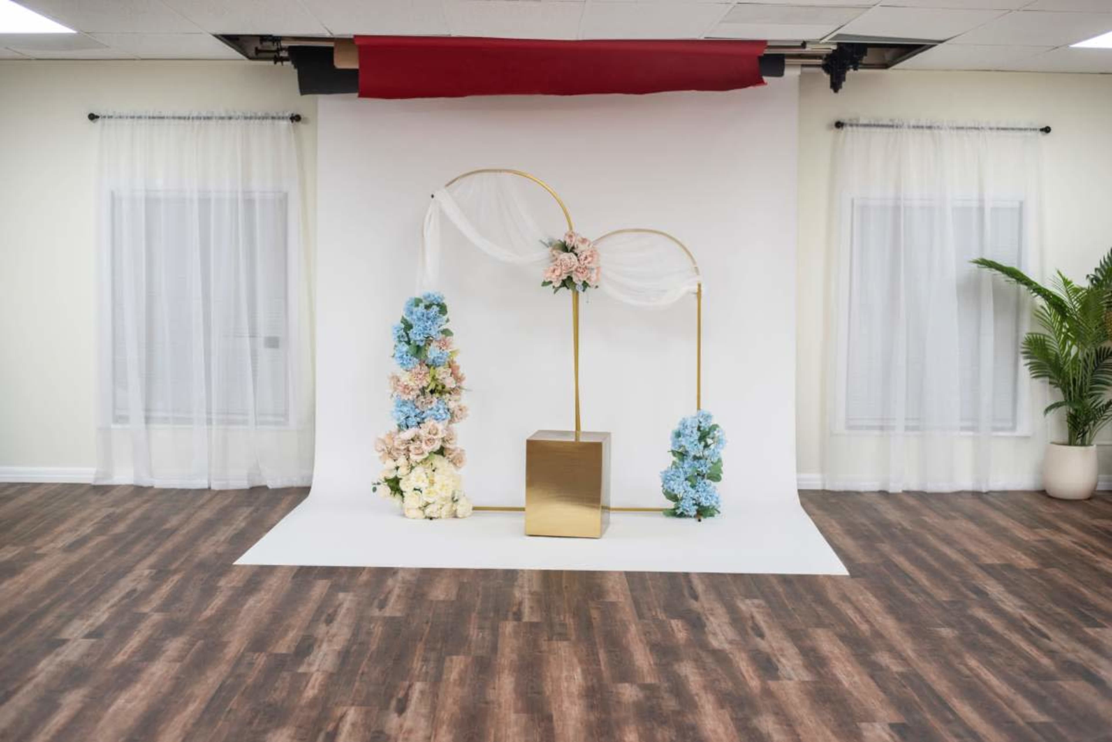The image shows a decorated photo backdrop featuring floral arrangements in pink and blue, draped fabric, and a gold cube pedestal, set against a white wall and wooden flooring.