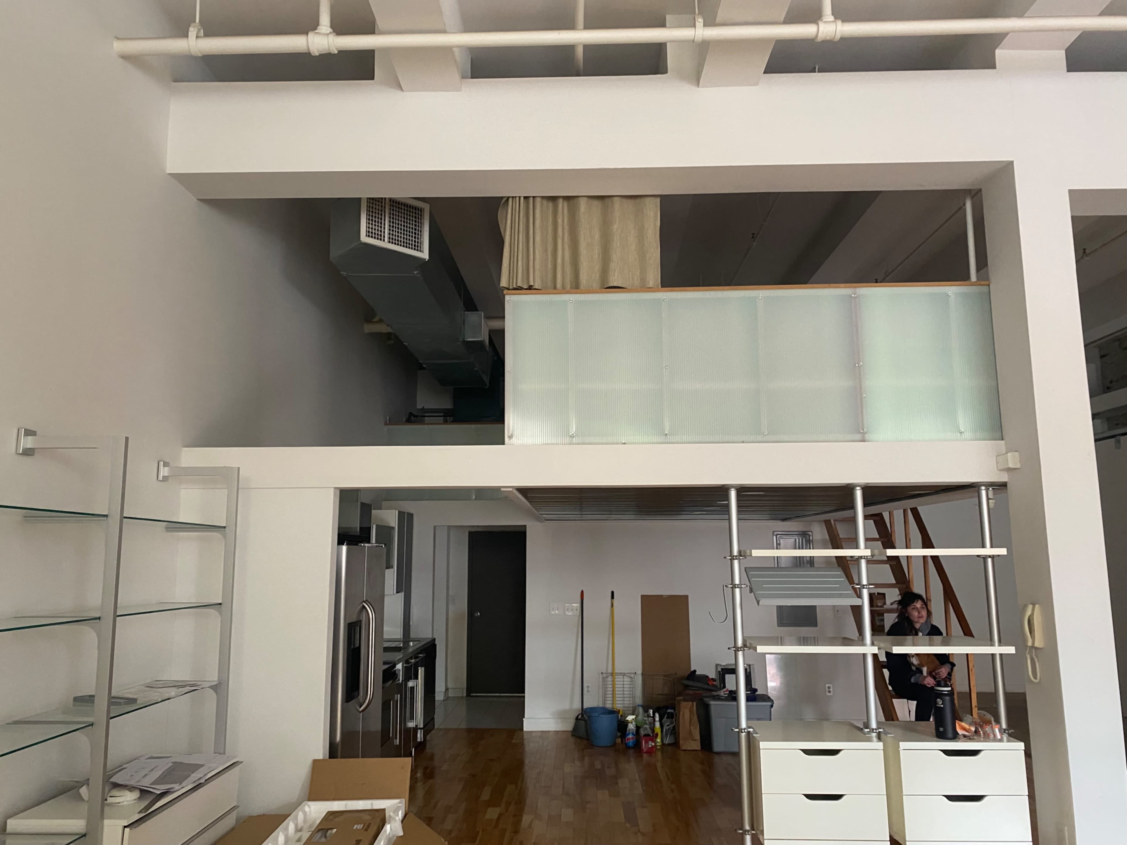 The image shows a spacious, unfinished interior of a loft-style apartment with a kitchen area and a raised platform featuring a railing.