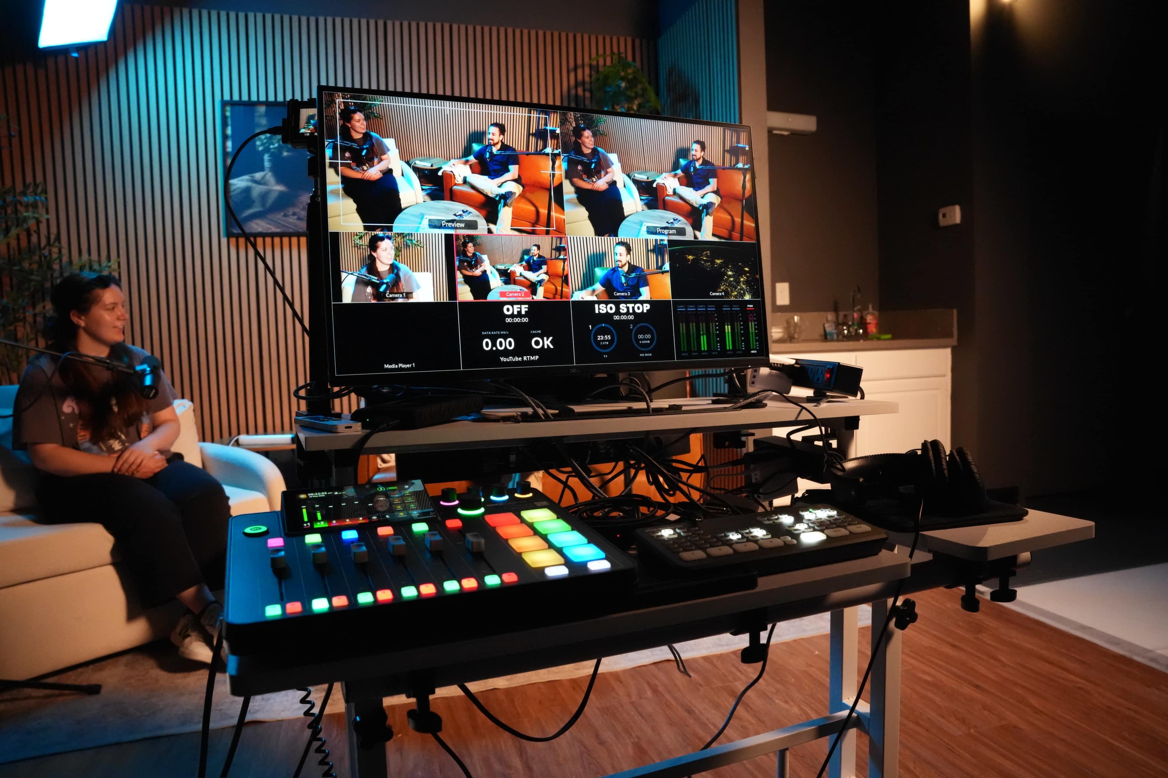 A control desk with a monitor displaying multiple camera feeds is set up in a studio, while a person sits on a couch in the background.
