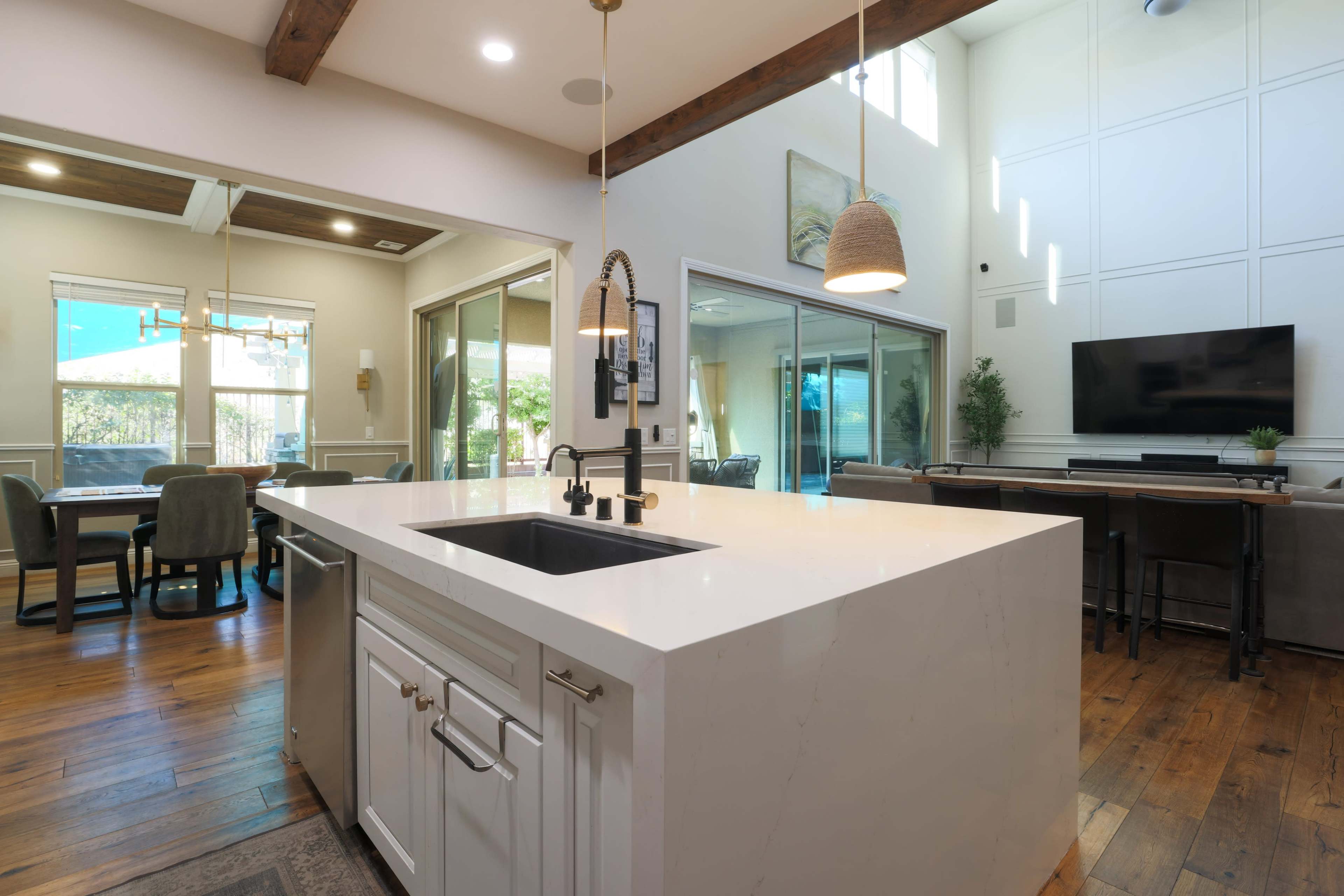 The image shows a modern kitchen with a large island, featuring a sink and stark white countertops, open to a dining area and living space with large windows and a television.