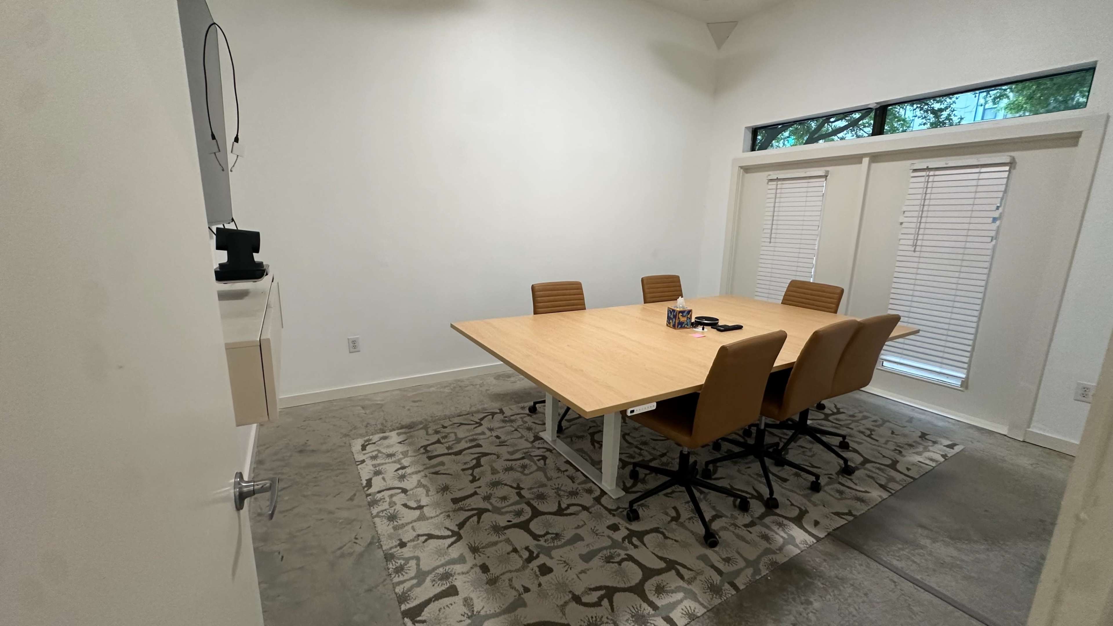 A large conference room features a rectangular table surrounded by six brown chairs, with a rug on the floor and a window allowing natural light.