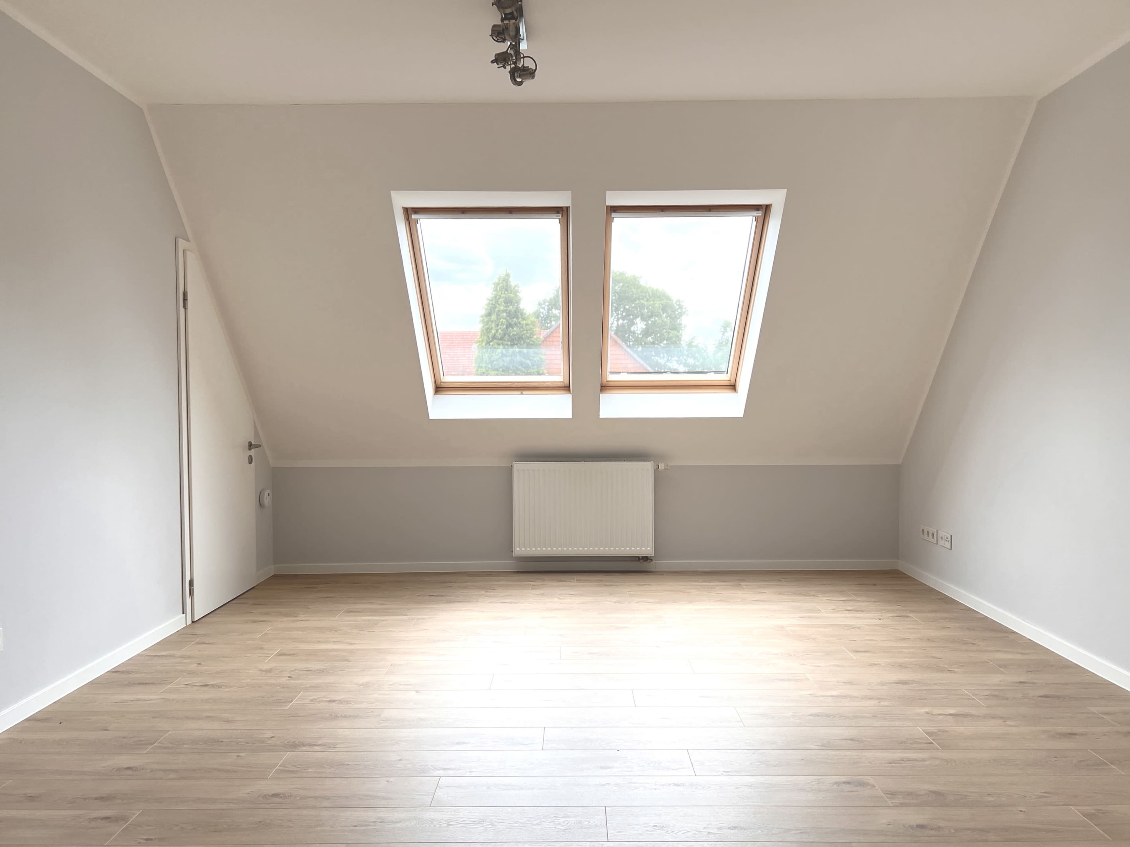 The image shows a spacious, empty room with two large skylights, wooden flooring, and a central heating radiator on the wall.