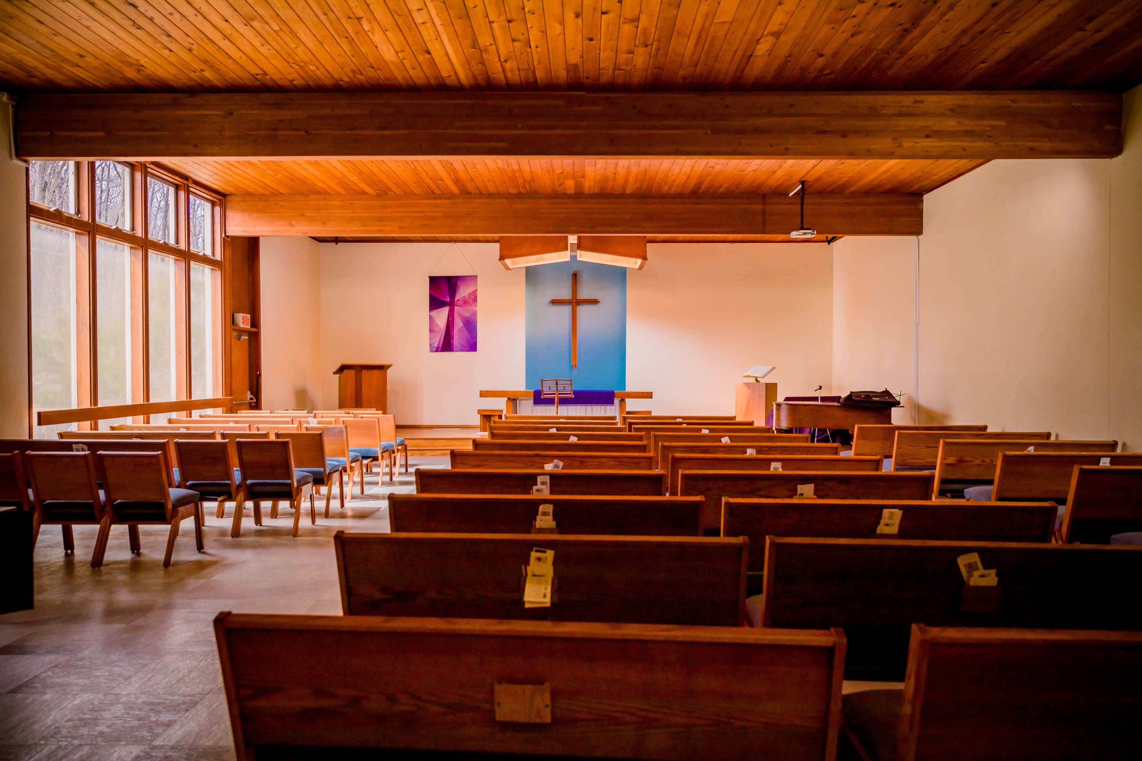 The interior of a church with wooden benches facing a blue wall featuring a large cross.