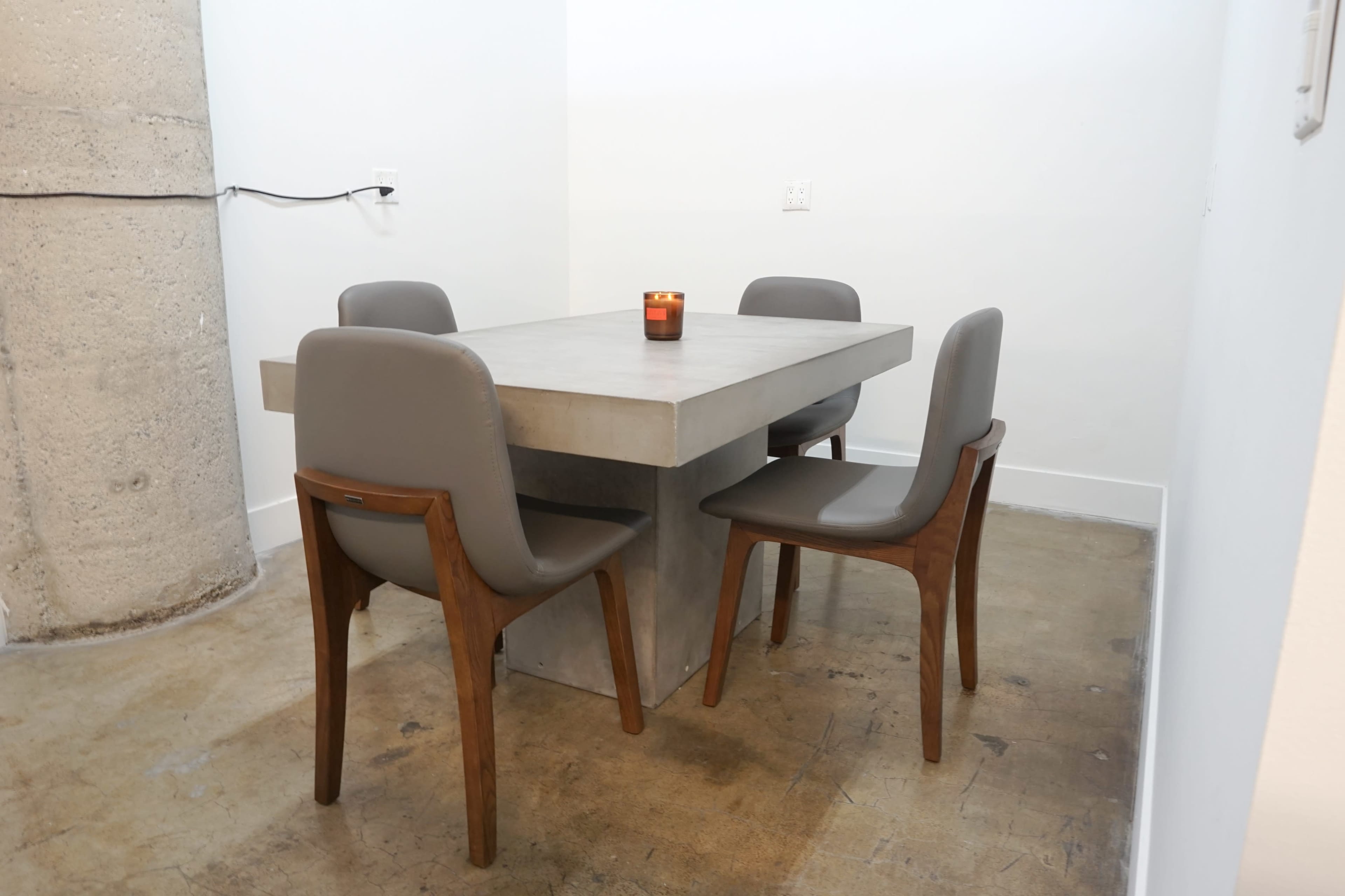 A concrete table with a simple design is surrounded by four wooden and upholstered chairs in a well-lit, minimalistic space.