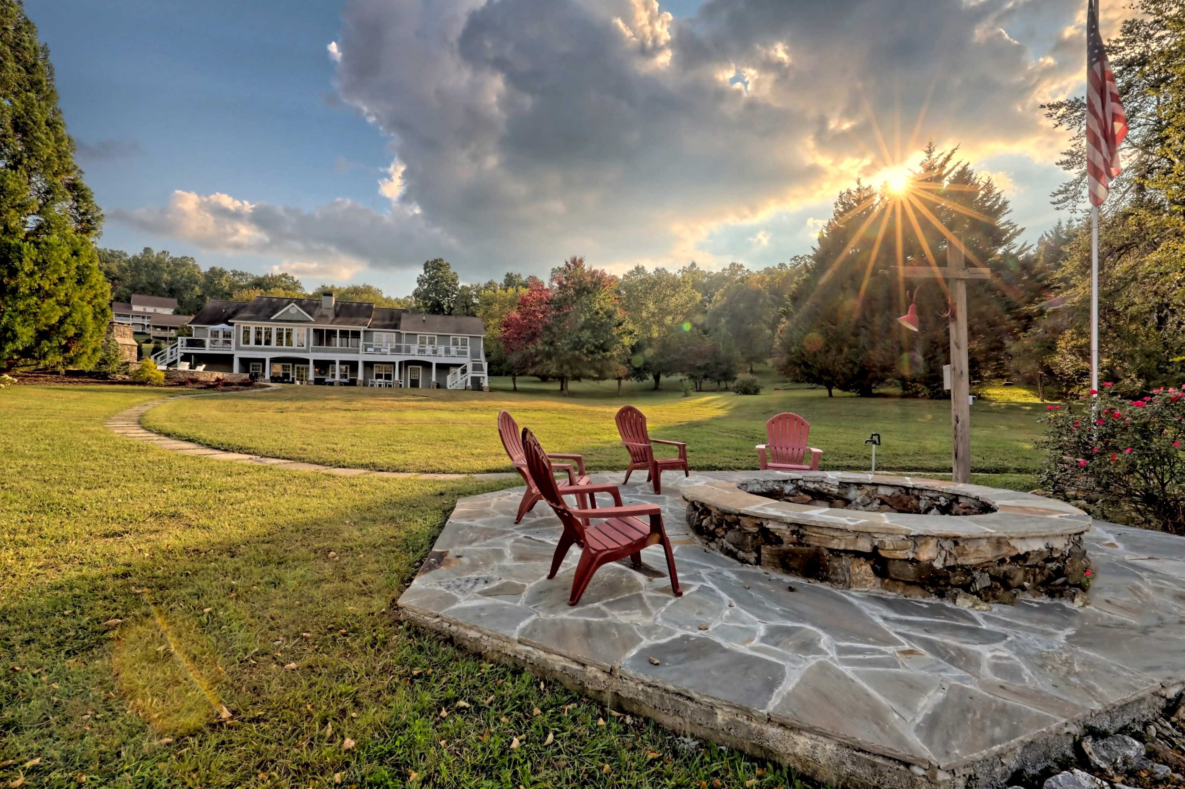 A landscaped backyard features a stone fire pit surrounded by red chairs, with a house in the background and a flagpole nearby.