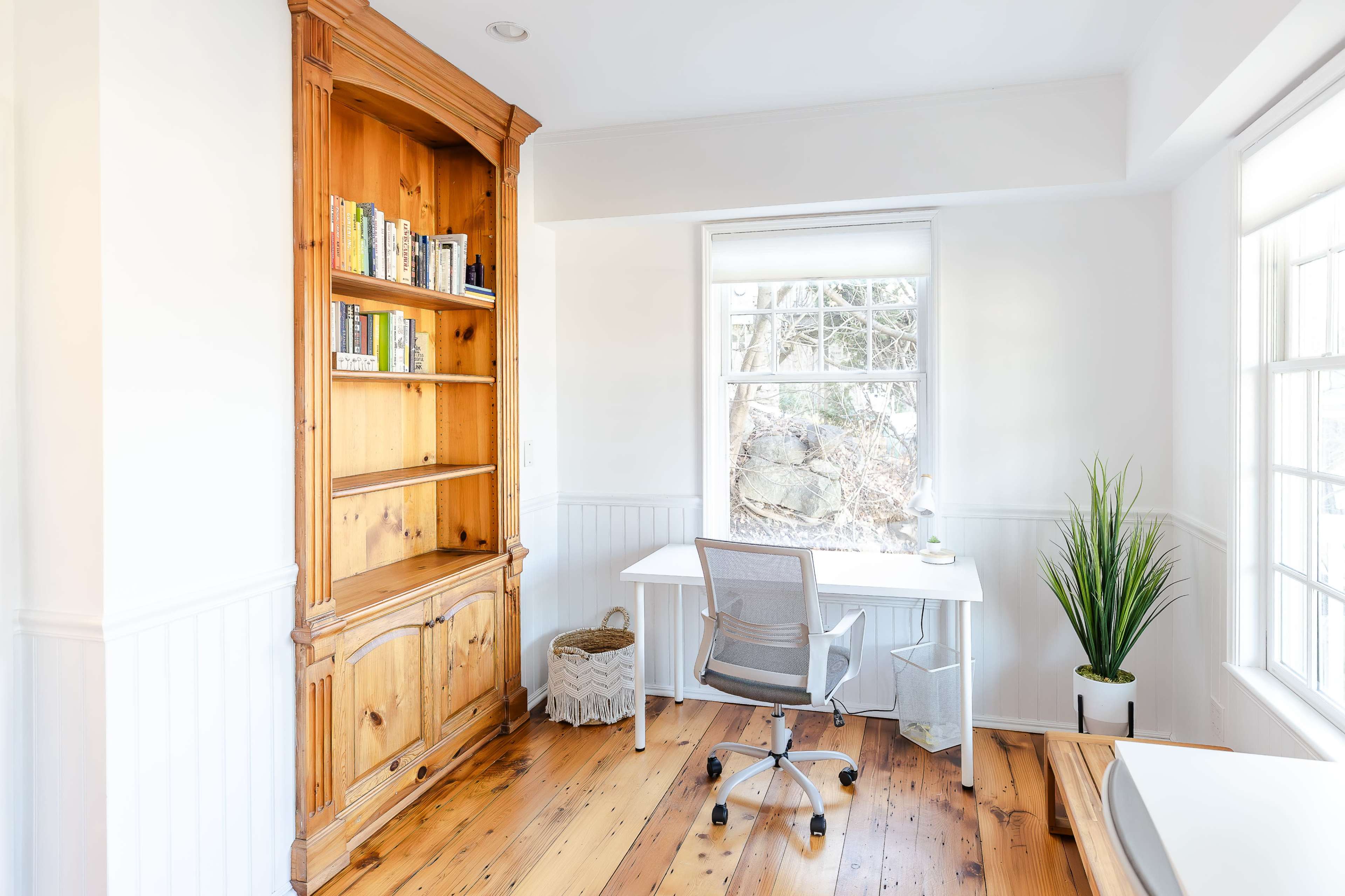 A bright, minimalistic home office featuring a wooden bookshelf, a white desk and chair, and a tall plant near large windows.