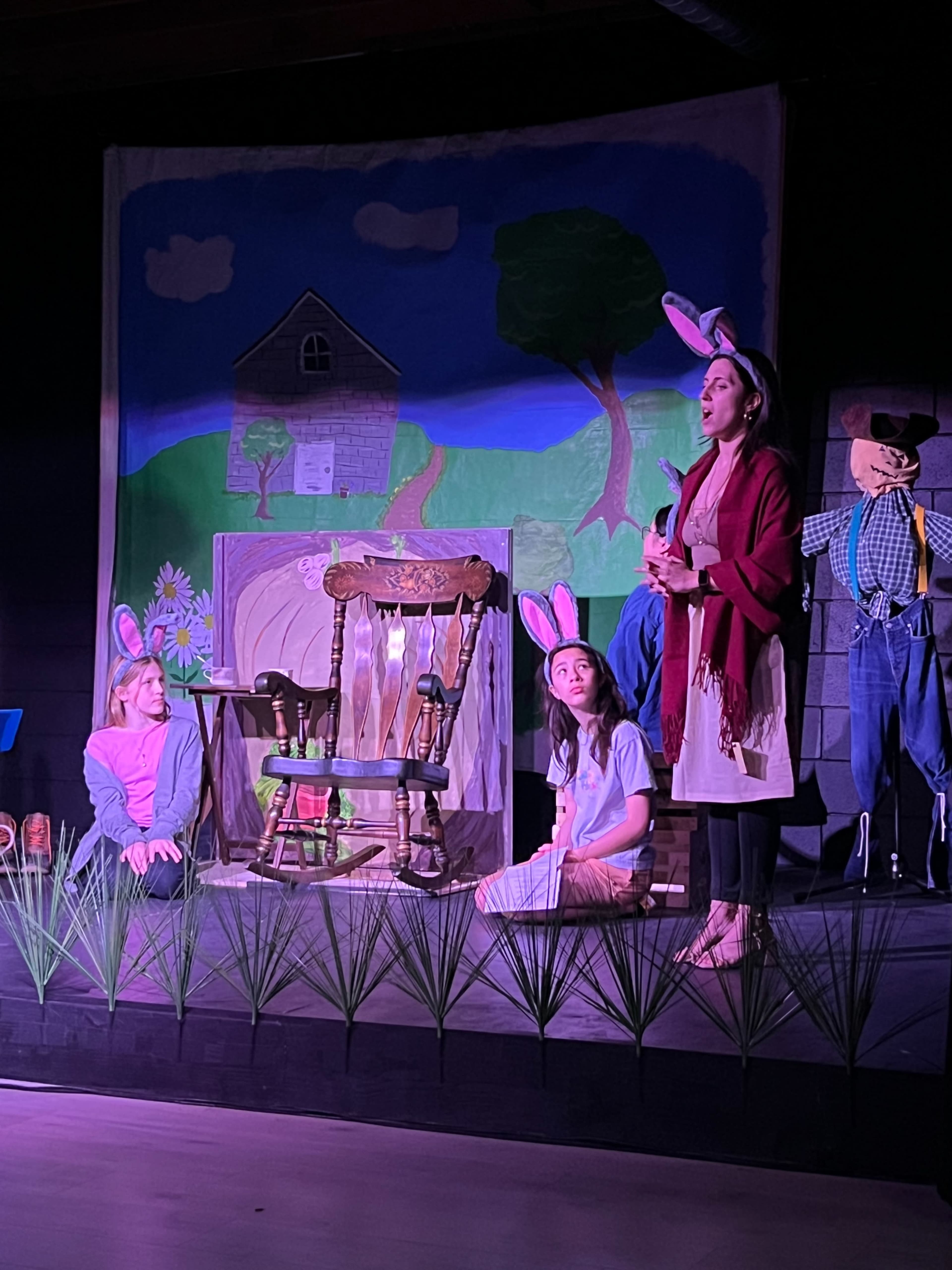 A stage setting features three people, two seated on the floor and one standing, all wearing bunny ears, with a painted backdrop of a house and tree.