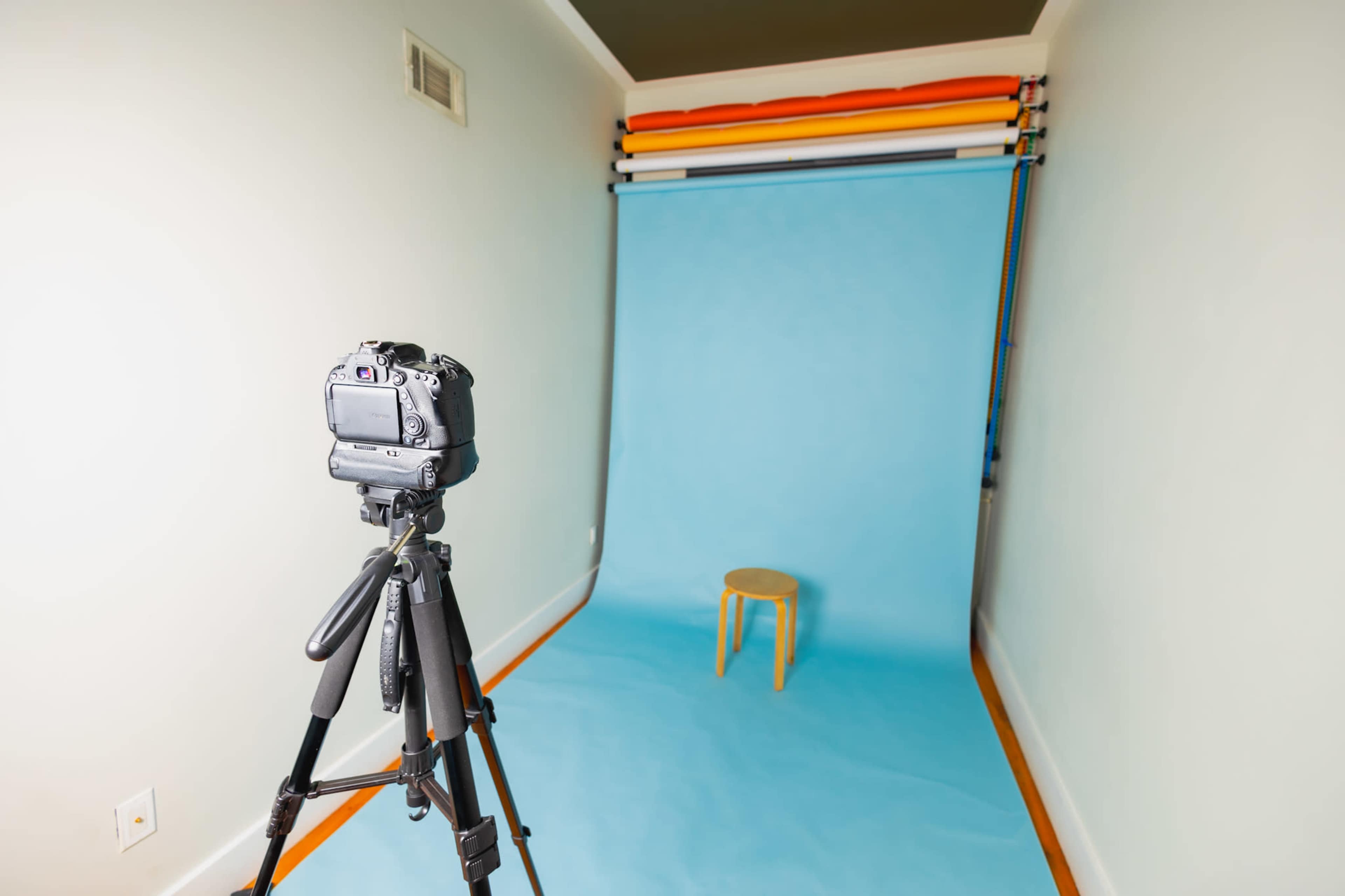 A camera on a tripod faces a plain blue backdrop with a wooden stool set in the center.