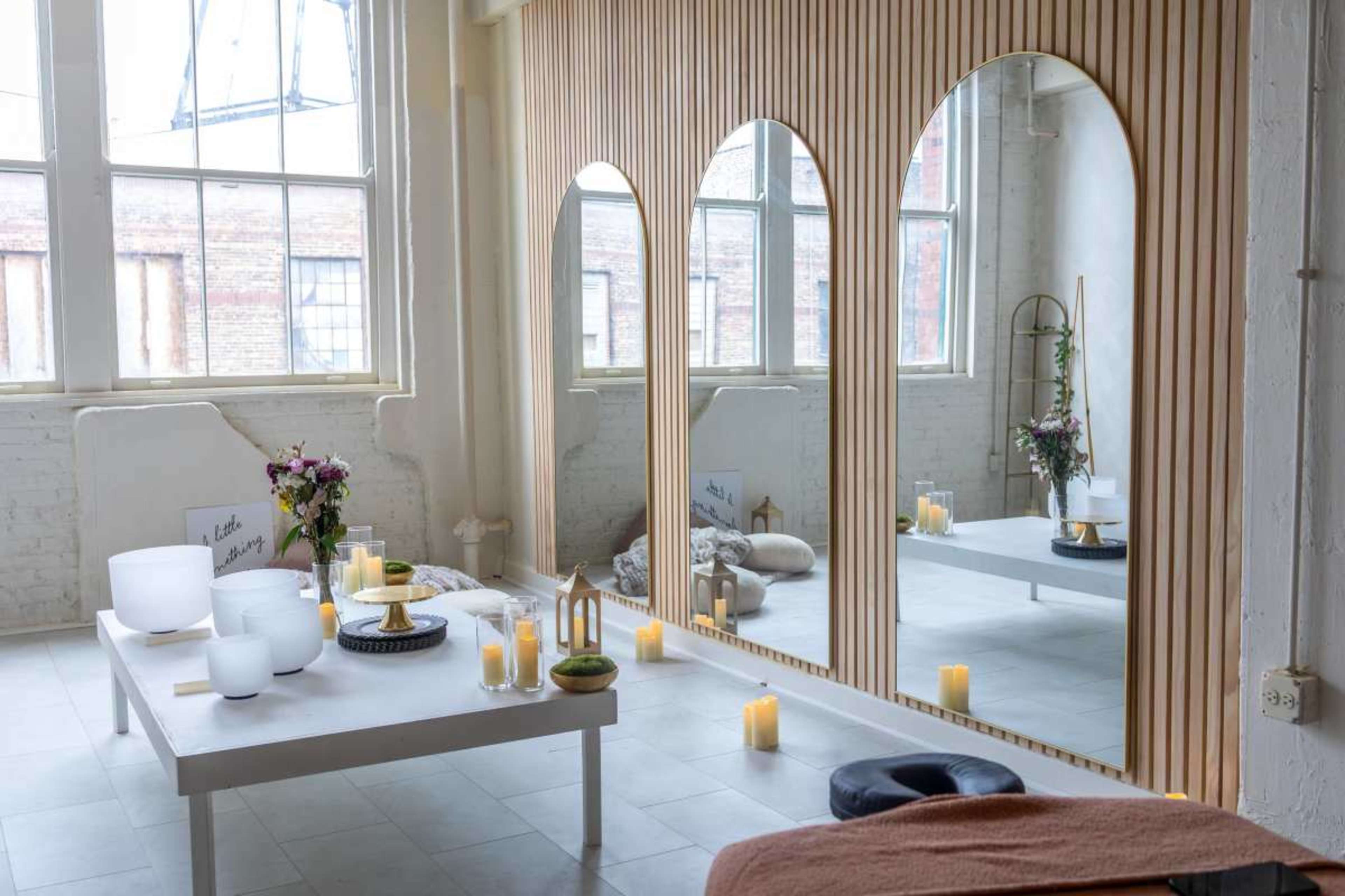 The image shows a bright, minimalist room with three large arched mirrors, a white table, candles, and floral arrangements.