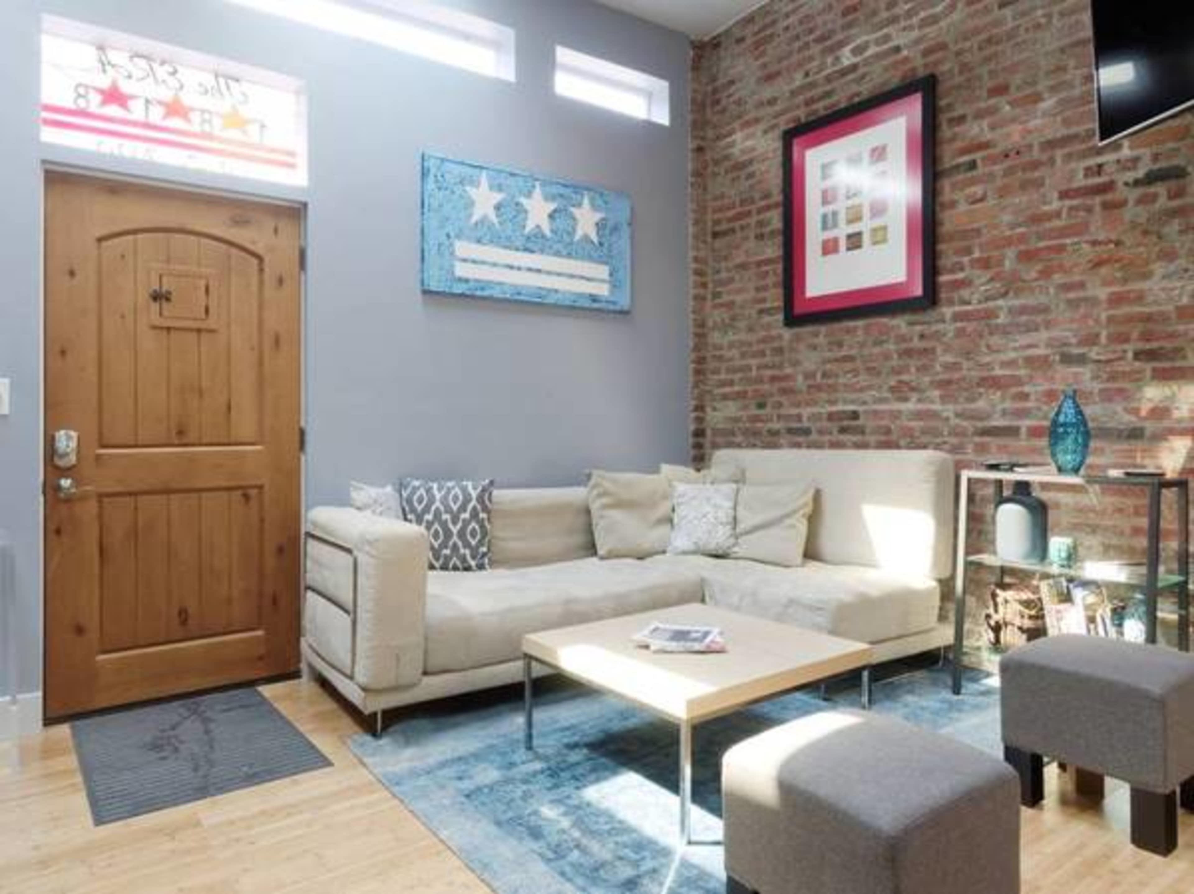 A modern living room with a beige sectional sofa, a wooden door, and a coffee table on a blue rug, framed by exposed brick walls.