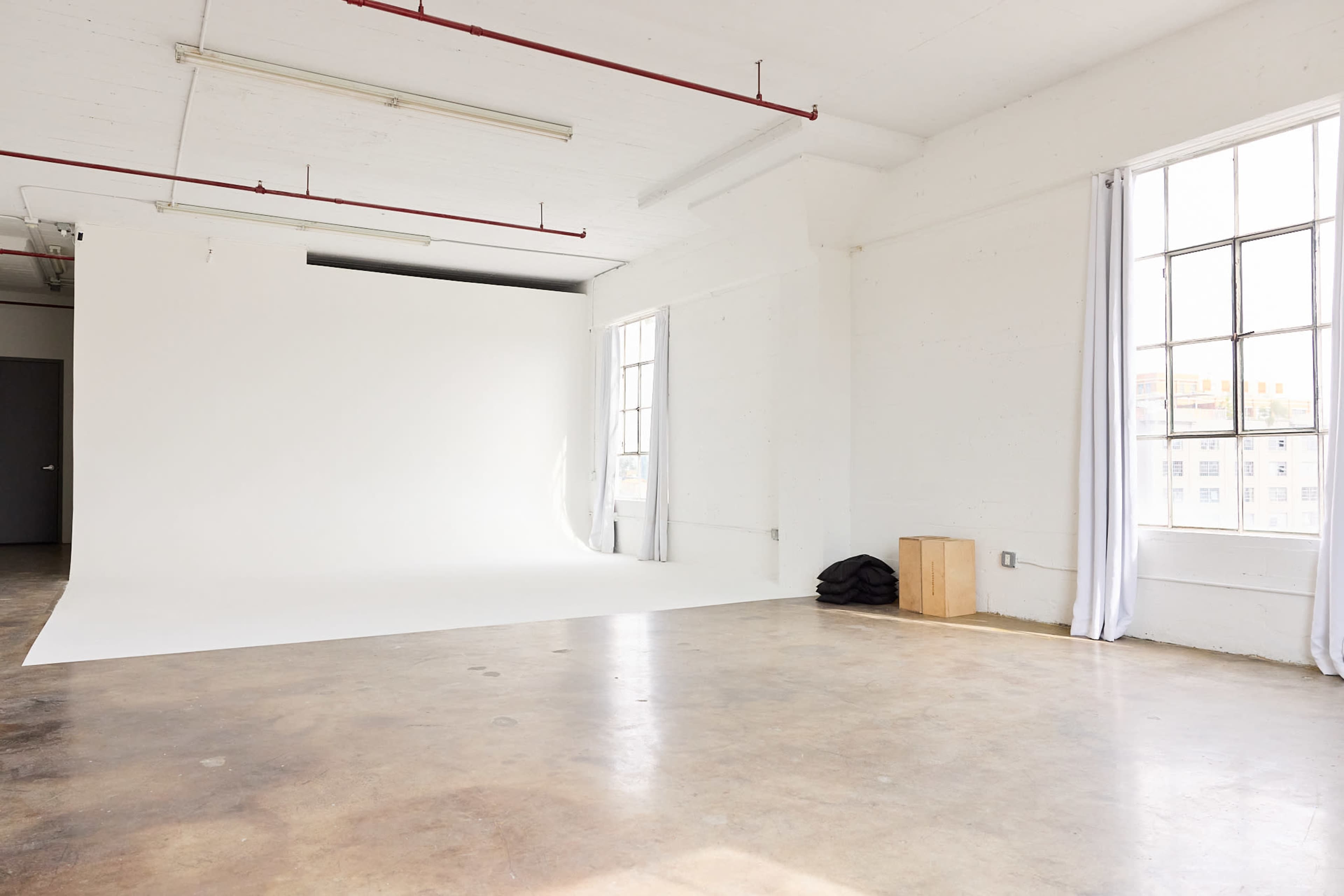 The image shows a spacious, well-lit photography studio with a white backdrop, two large windows, and a concrete floor.