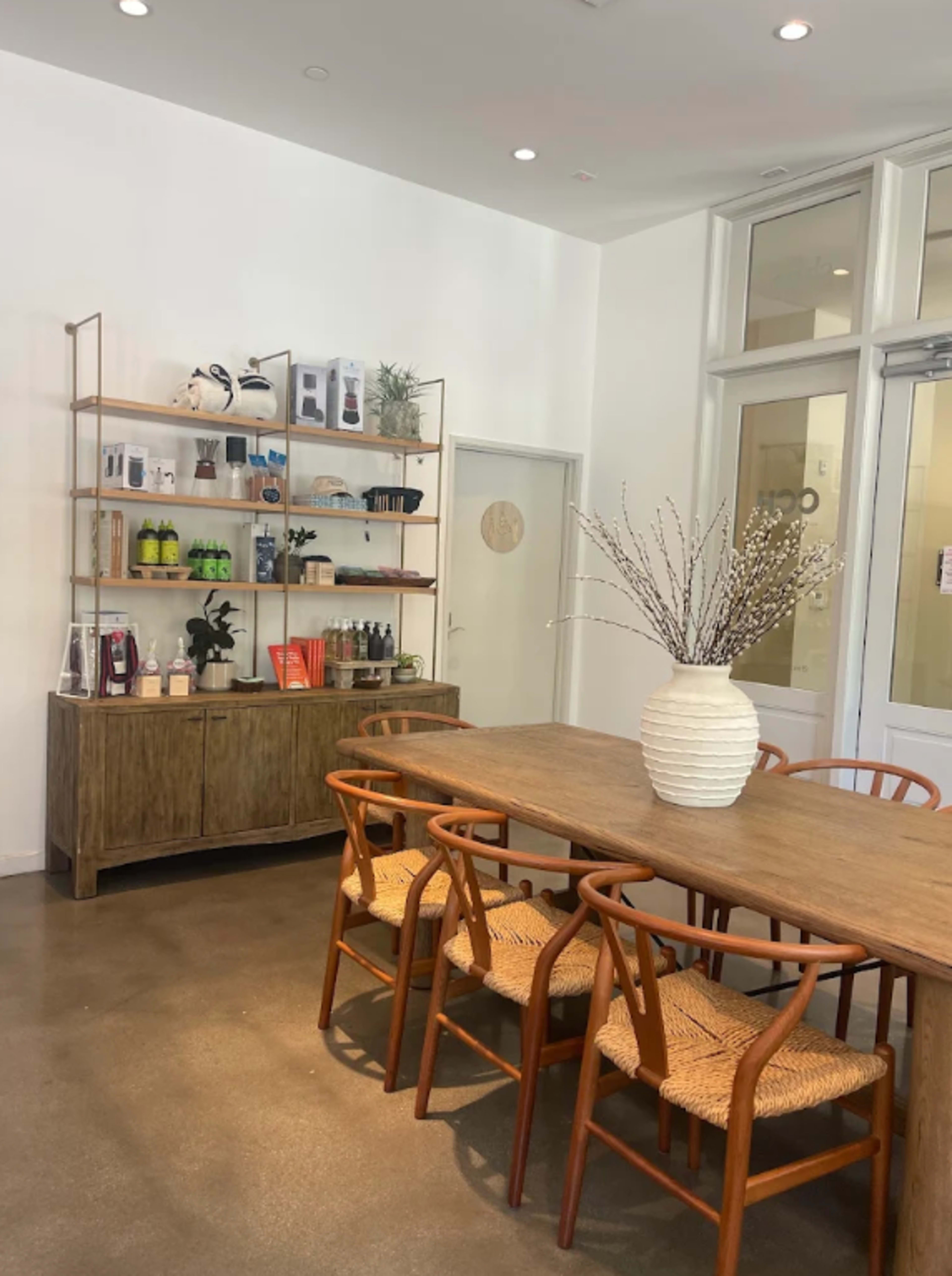 The image shows a clean, modern interior of a room featuring a wooden table with chairs, a display shelf filled with products, and a vase on the table.