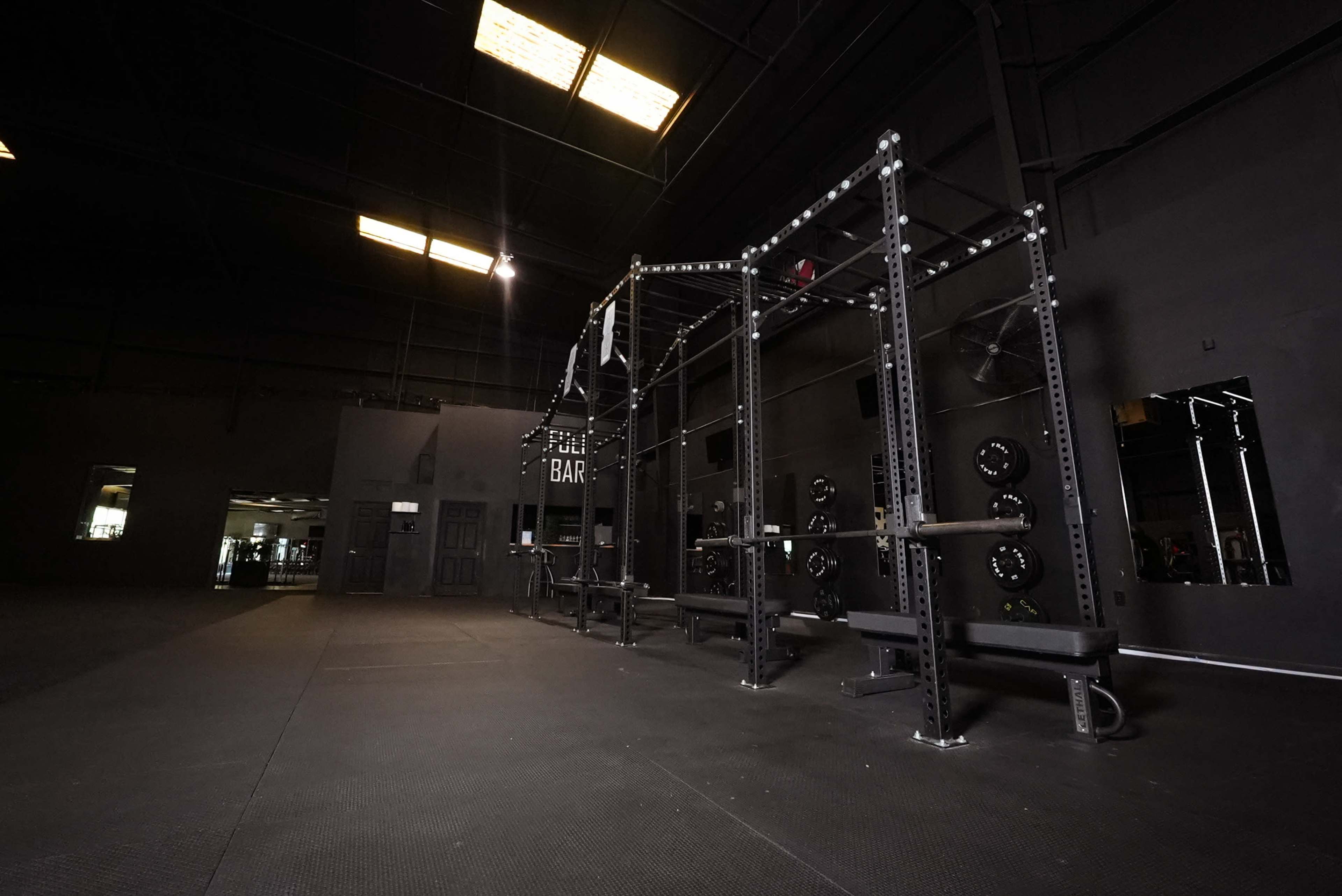The image shows a spacious gym interior featuring a black workout rig with benches and weight plates, under a high ceiling with natural light filtering through skylights.