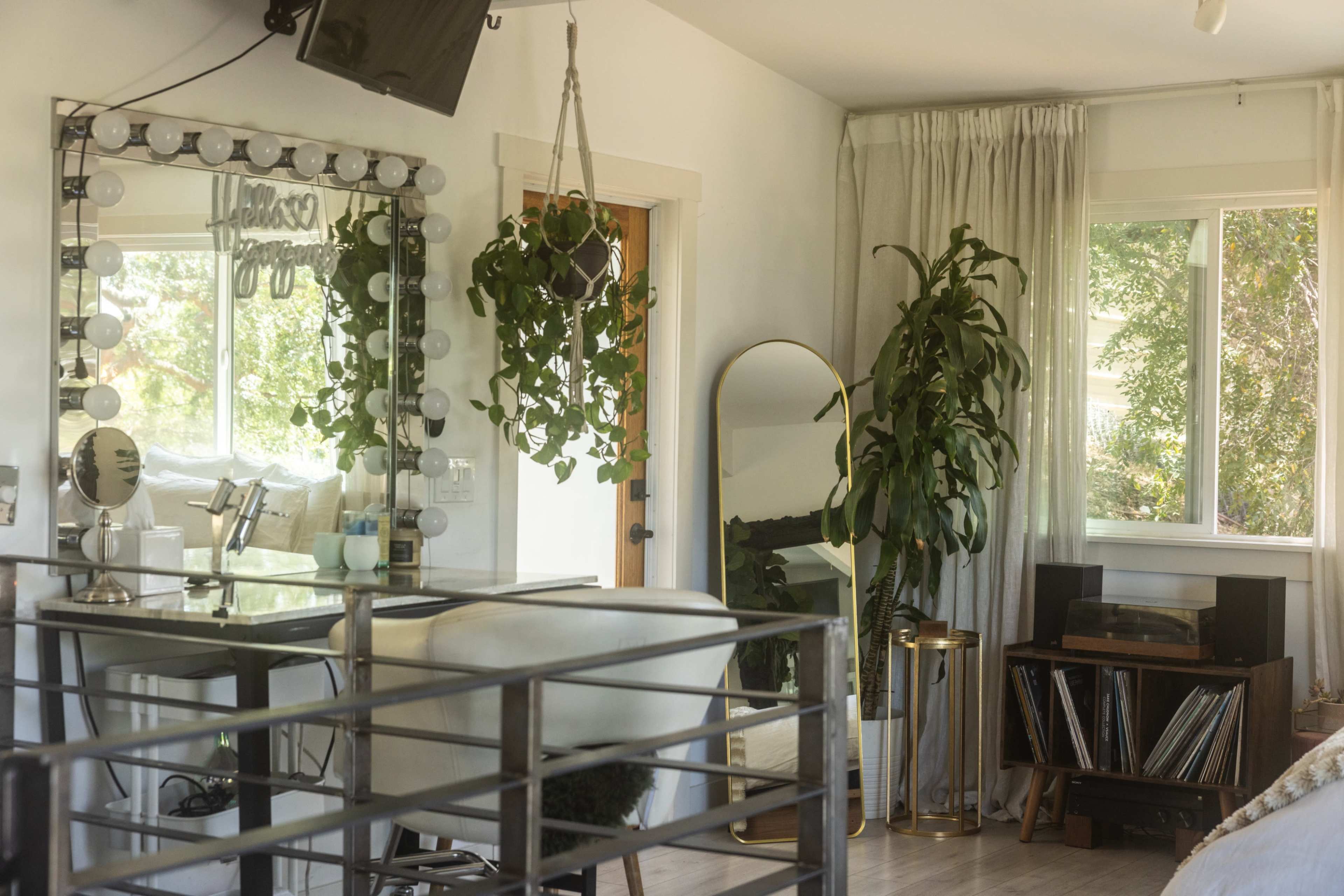 The image shows a bright, modern room with a makeup vanity, a large mirror with lights, a tall indoor plant, and a record player beside a collection of vinyl records.