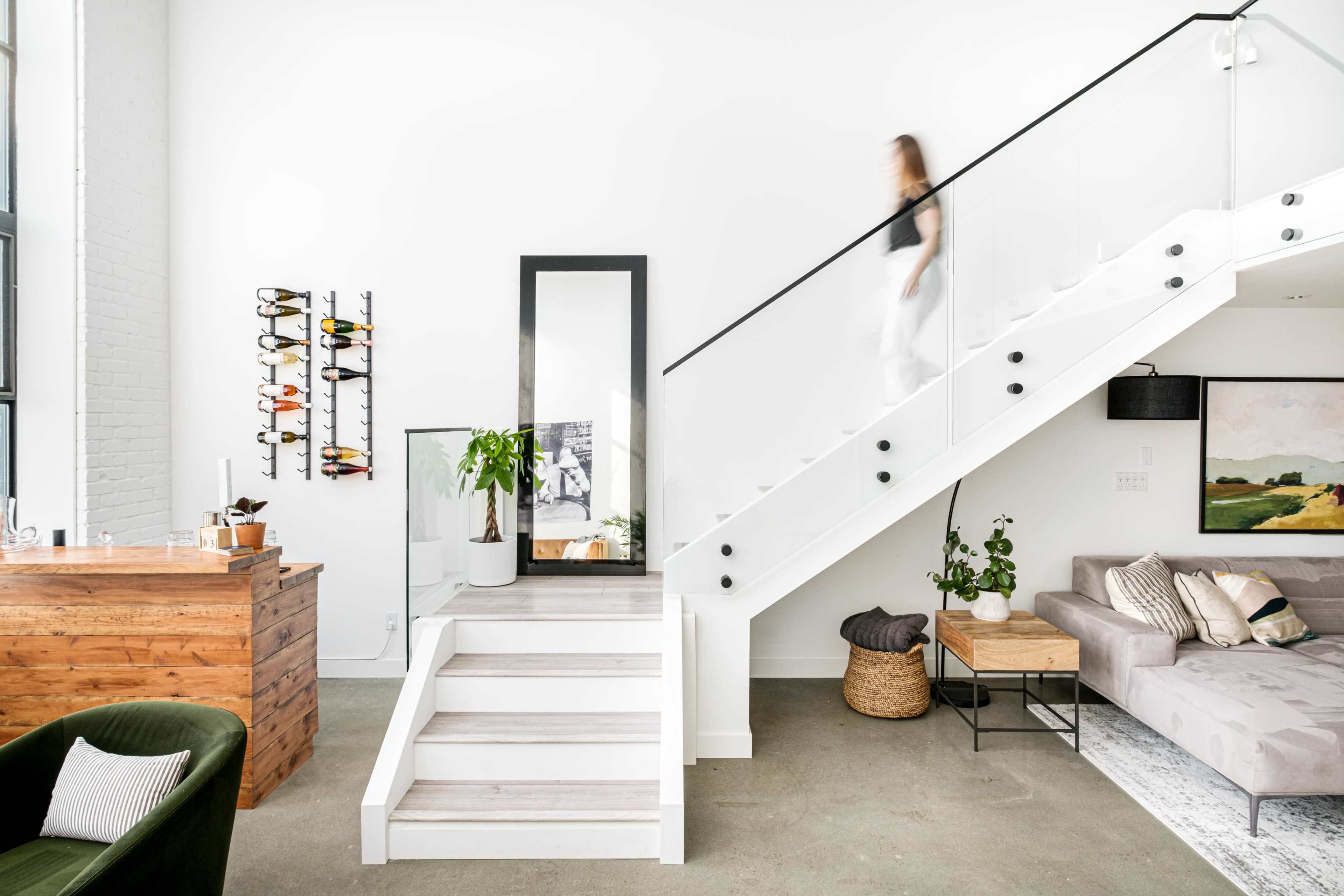 A woman walks up a staircase next to a living area featuring a sofa, a plant, and a wooden bar.