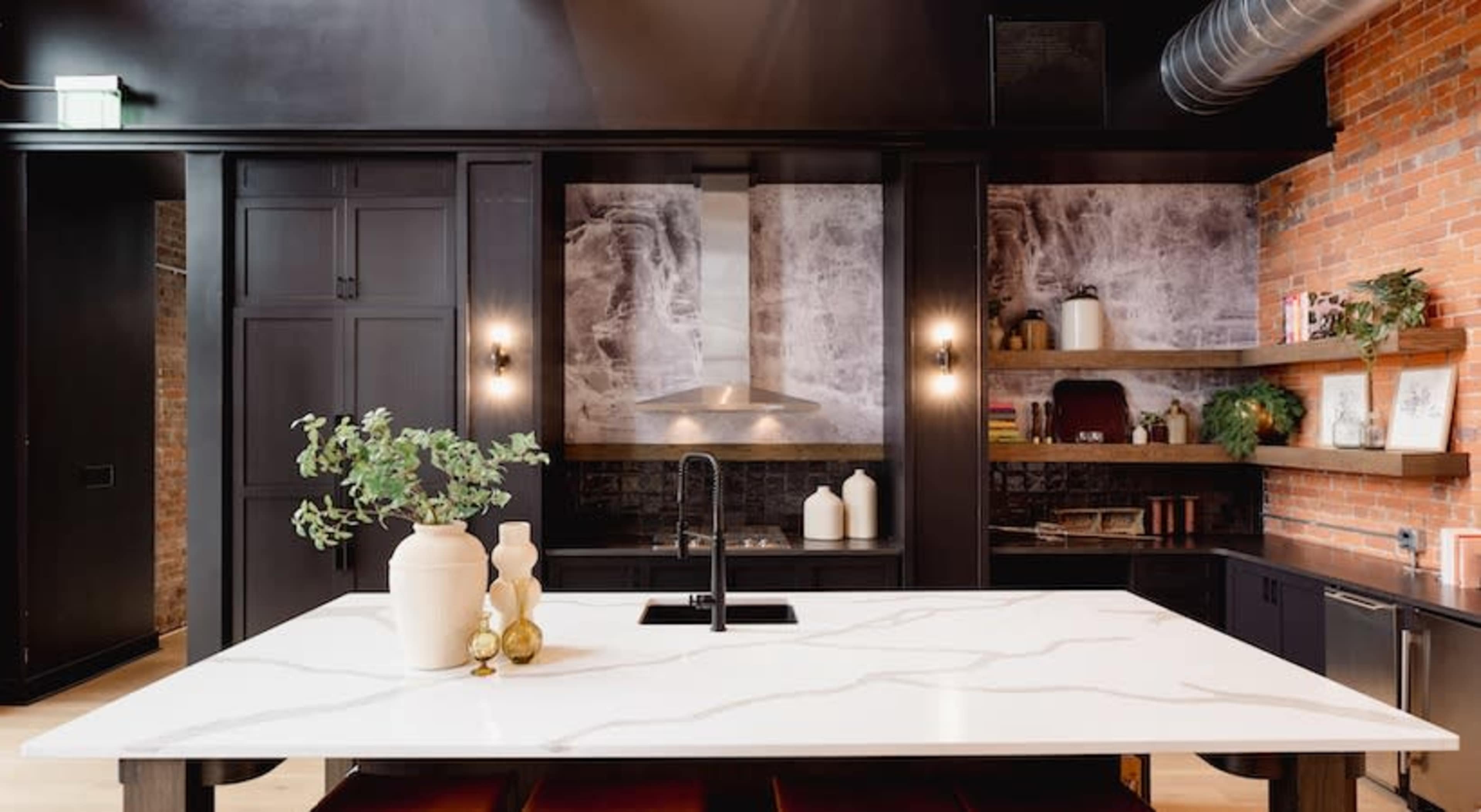 The image shows a modern kitchen with a large marble island, dark cabinetry, and textured wall panels, all complemented by decorative shelving.
