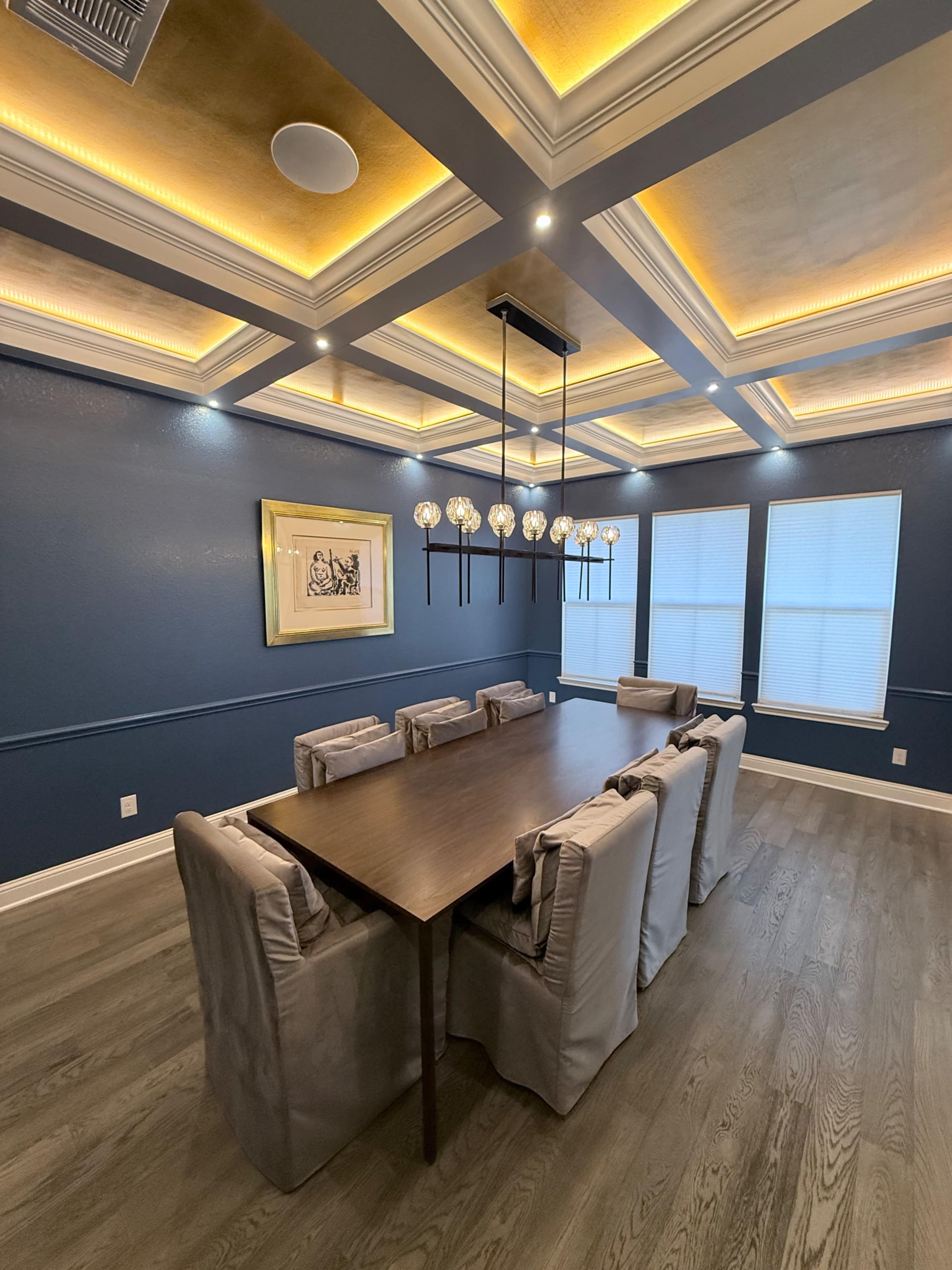 The image shows a modern dining room with a large wooden table surrounded by upholstered chairs, featuring a coffered ceiling with ambient lighting and three windows.