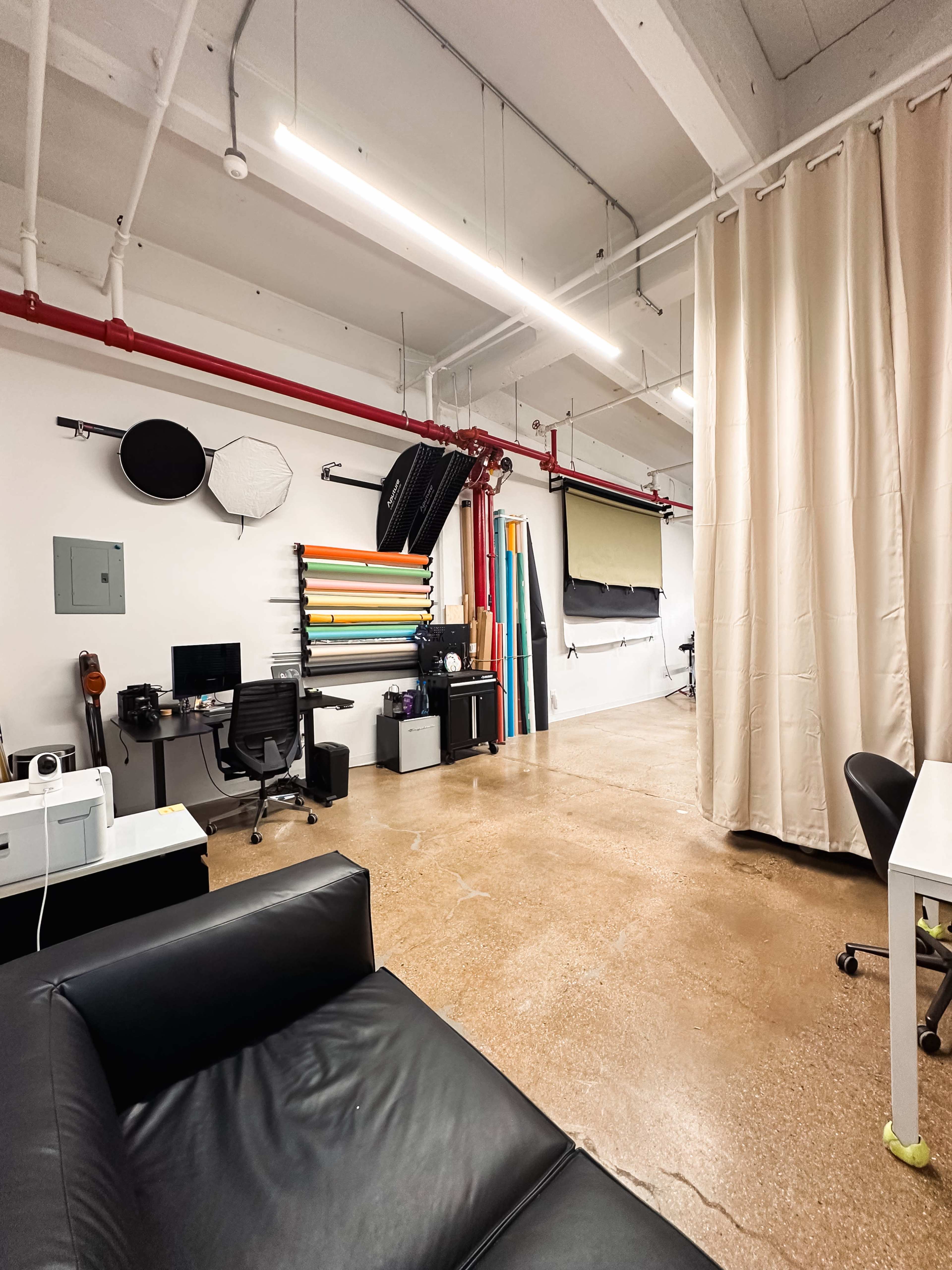 A spacious studio interior with various photography backdrops hanging on the wall, a desk with a computer setup, and a black couch in the foreground.