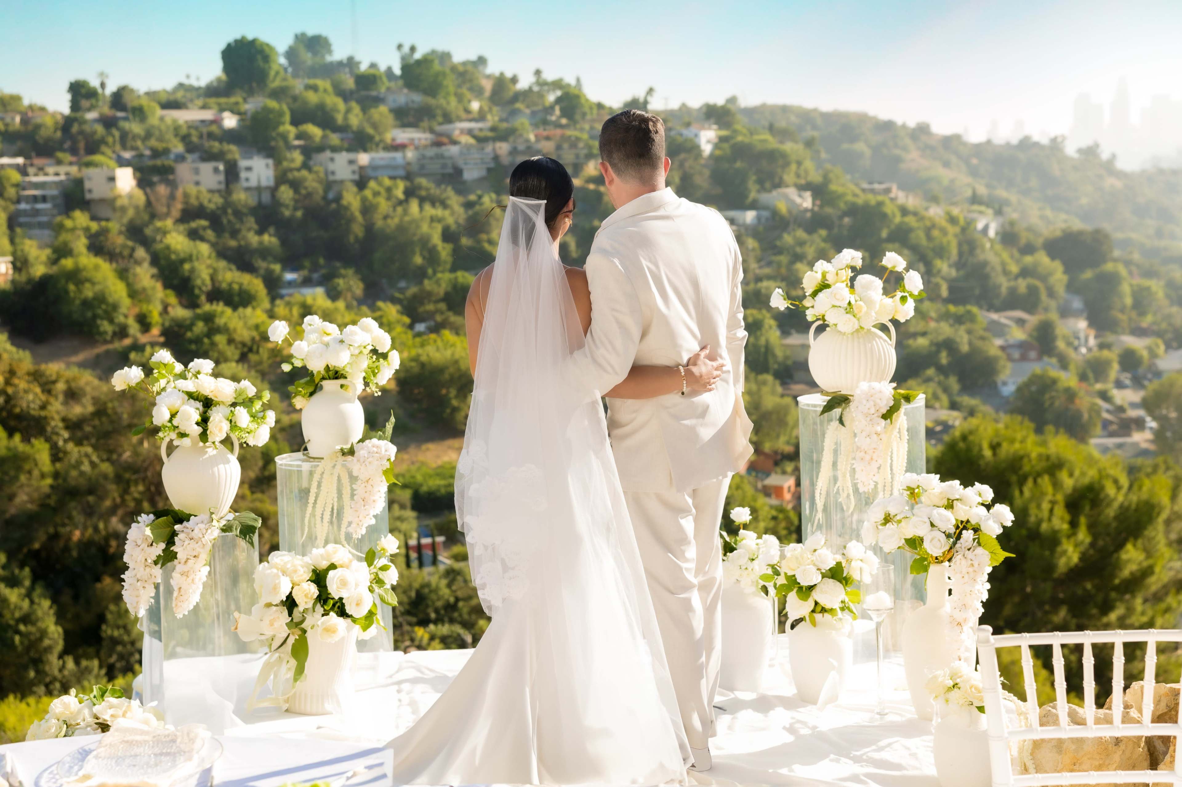 A couple stands together in wedding attire, facing a scenic view of hills and cityscape, surrounded by floral arrangements.