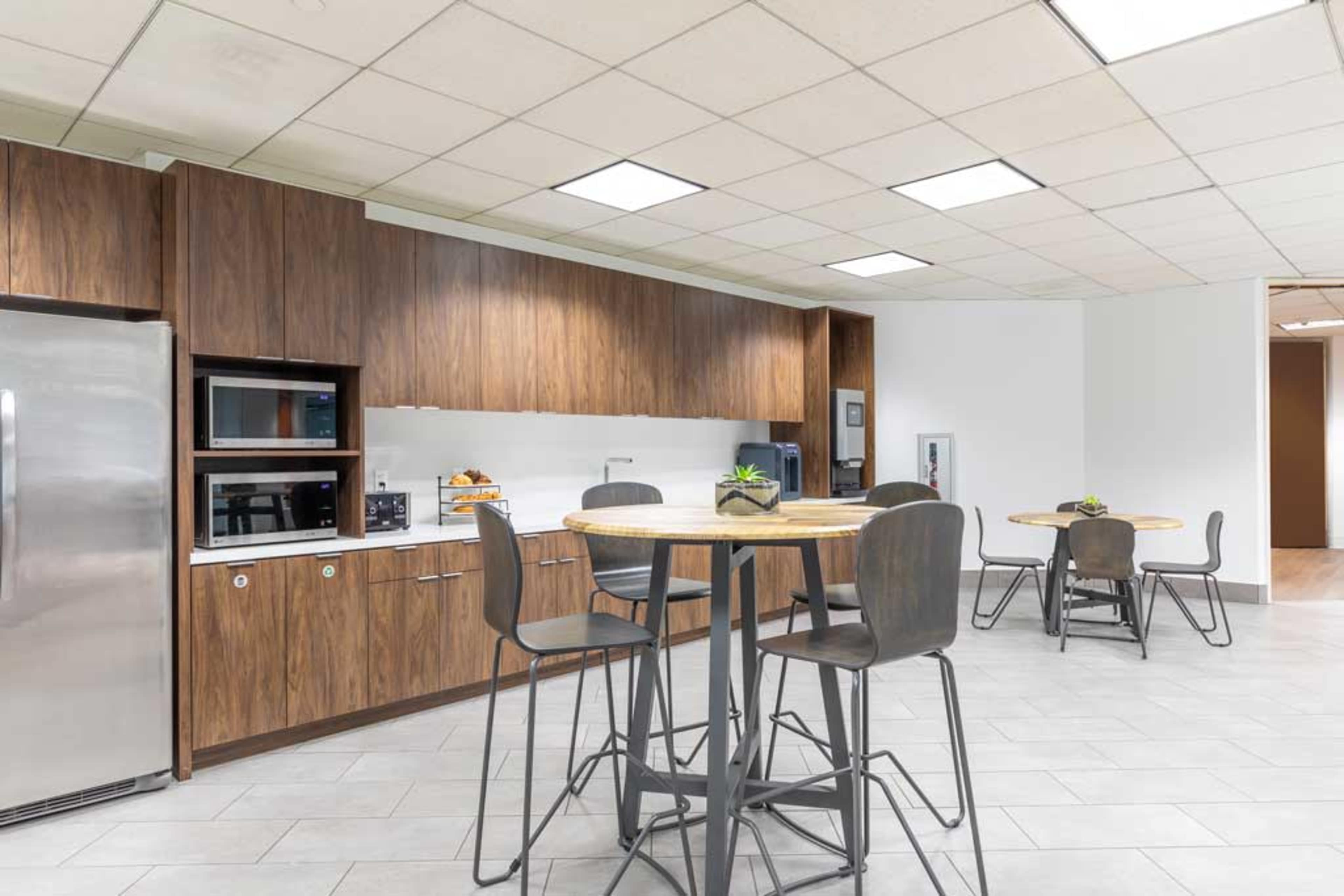 The image shows a modern kitchen area with wooden cabinetry, stainless steel appliances, and several round tables with high chairs.