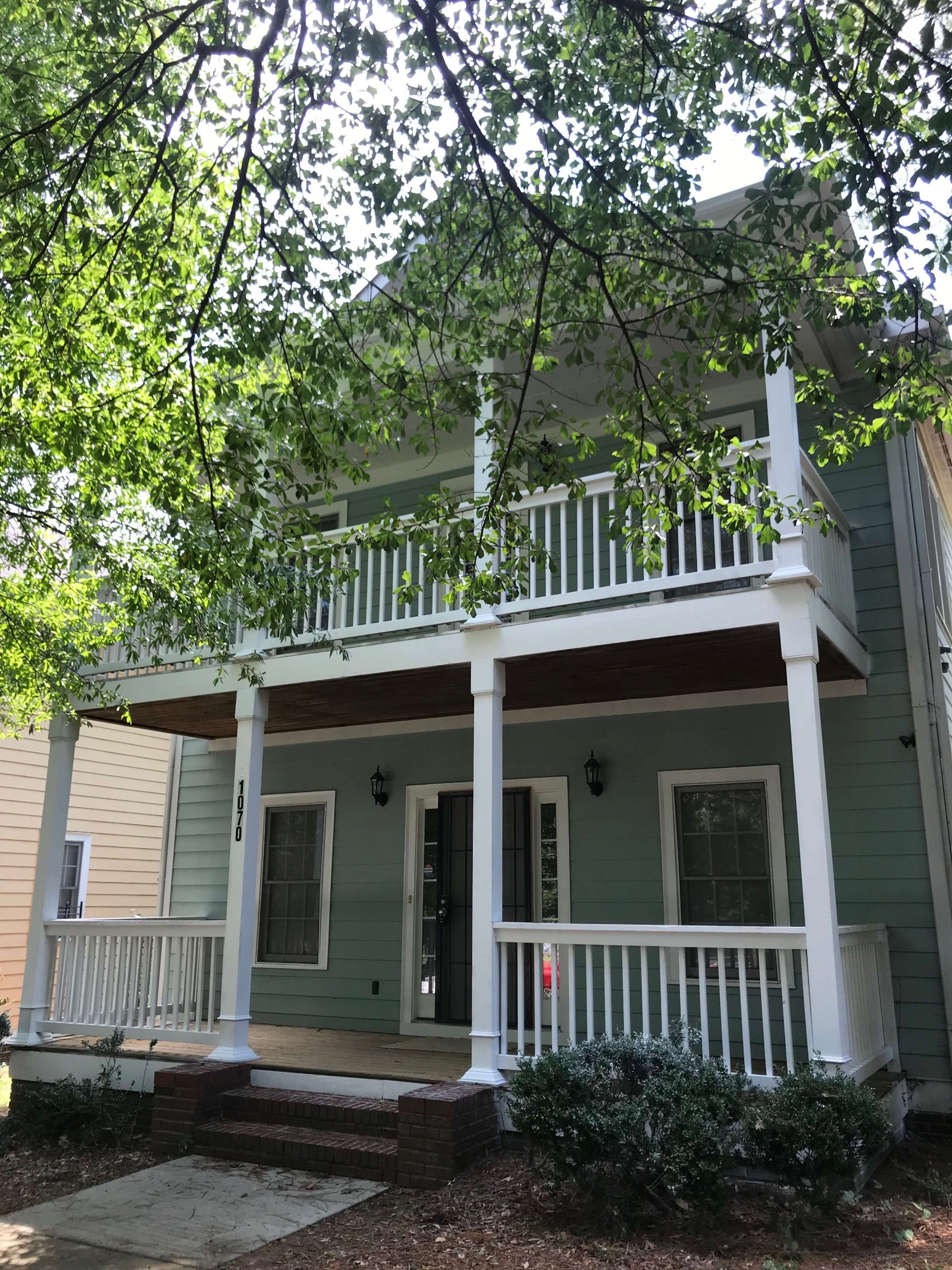 A two-story house with a light green exterior, a front porch supported by white columns, and a walkway leading to the entrance.