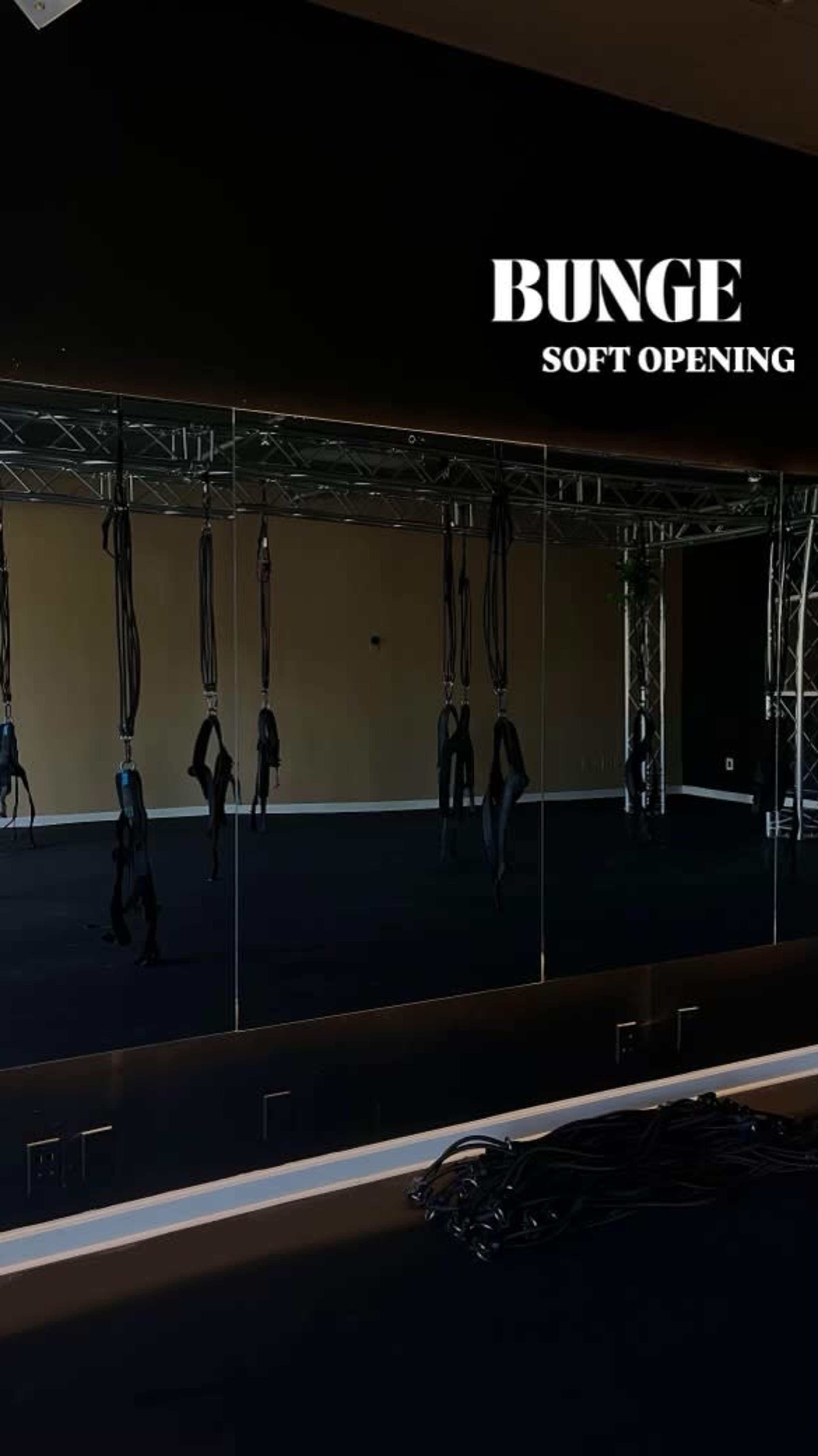 The image shows a fitness studio featuring a large mirror and suspended equipment against a black wall.