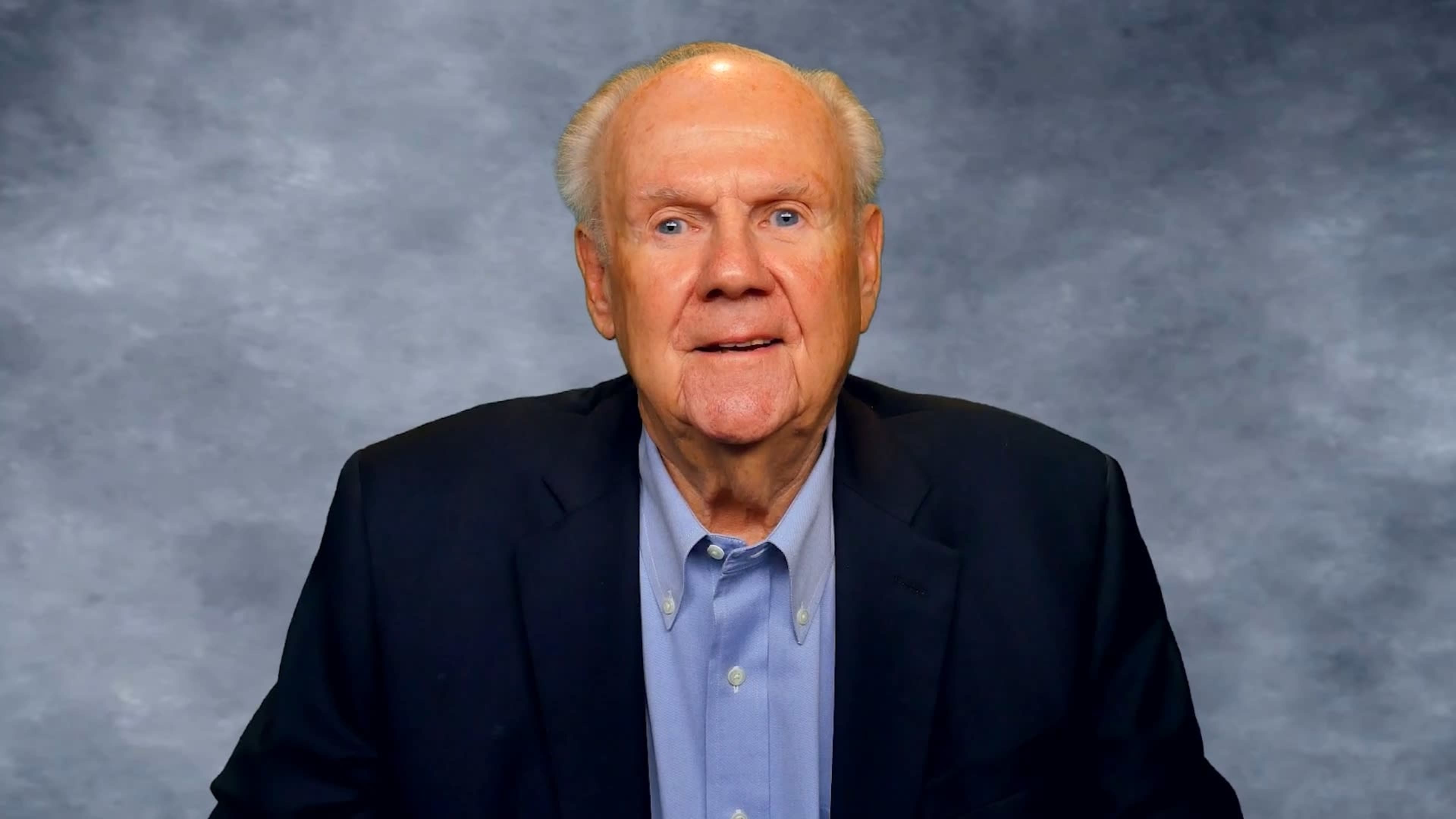 A man in a dark suit and light blue shirt sitting in front of a gray backdrop.