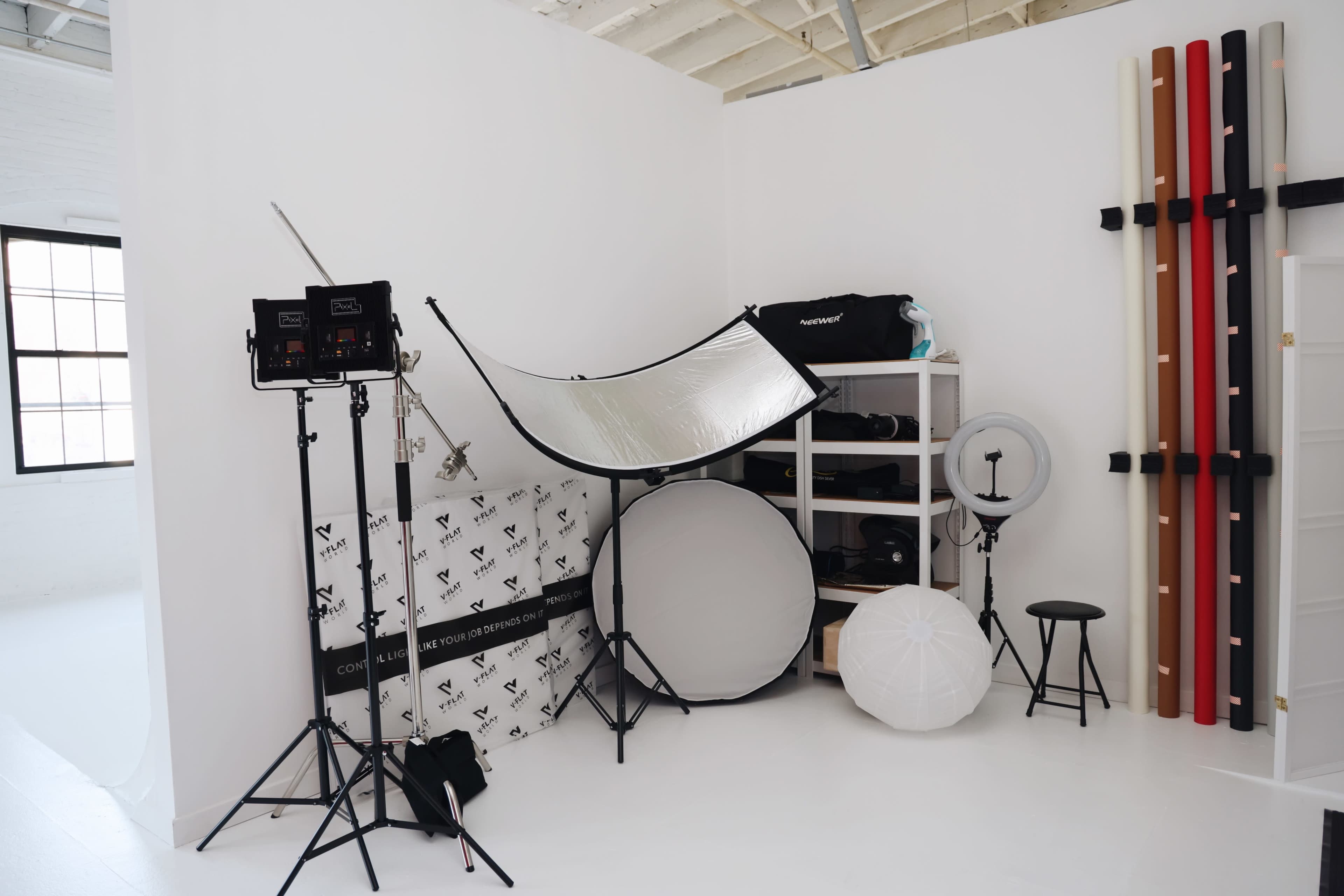 A photography studio setup featuring various lighting equipment, reflectors, and background support, all arranged against white walls.