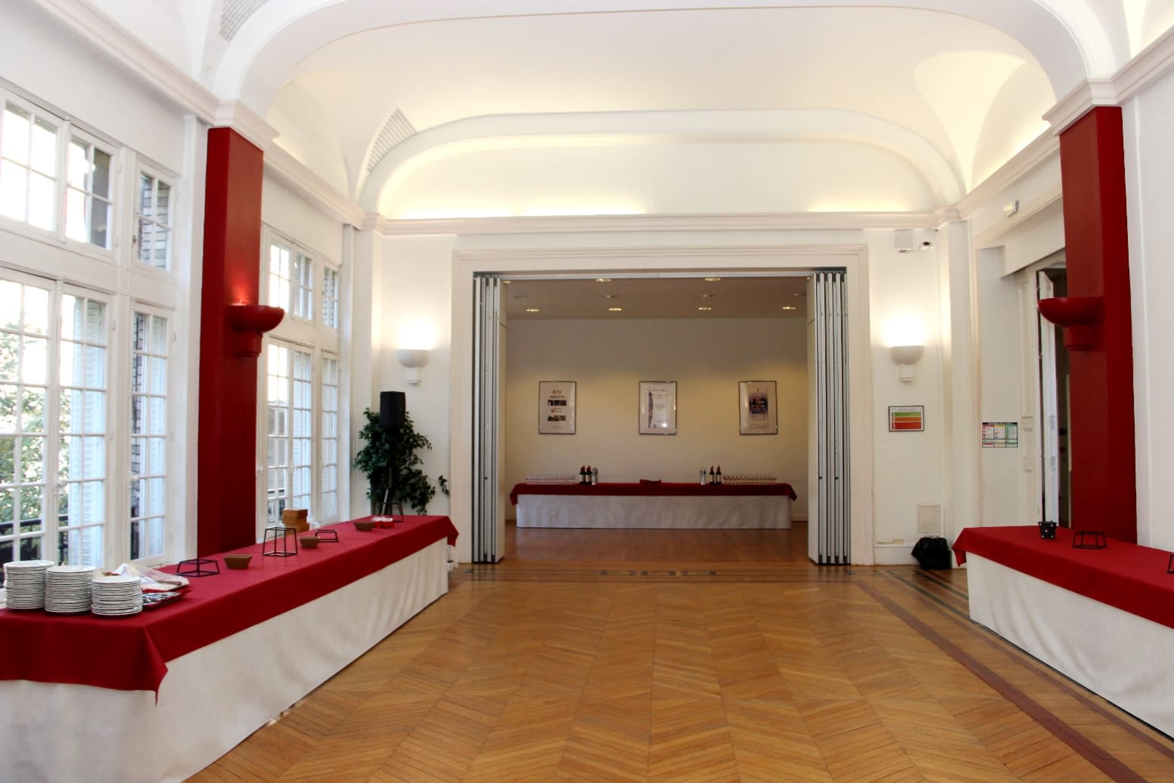 A spacious room with wooden flooring and white walls, featuring red table coverings along the sides and a set of sliding doors at the back.