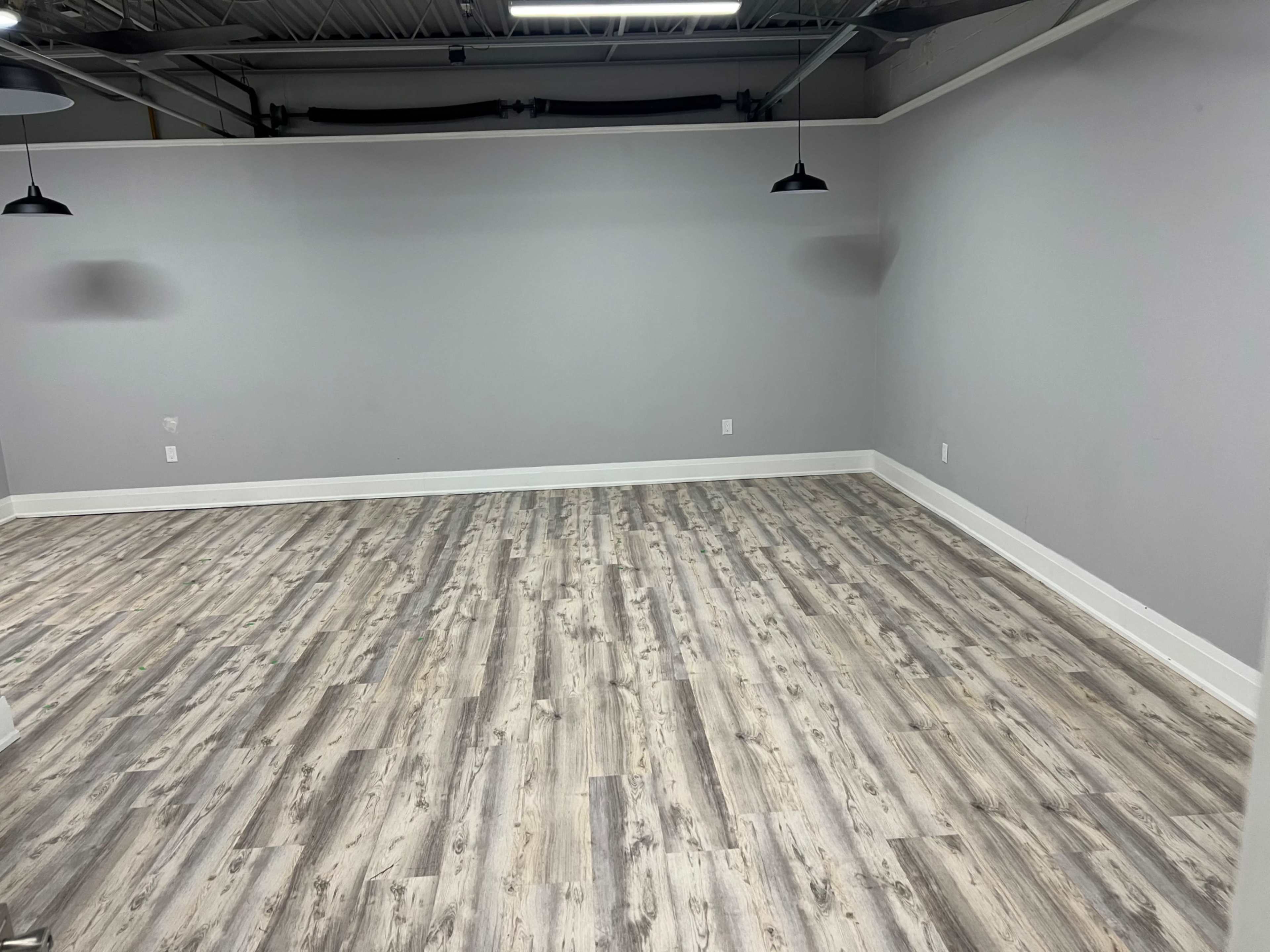 An empty room with gray walls and a wooden floor, featuring two pendant lights hanging from the ceiling.