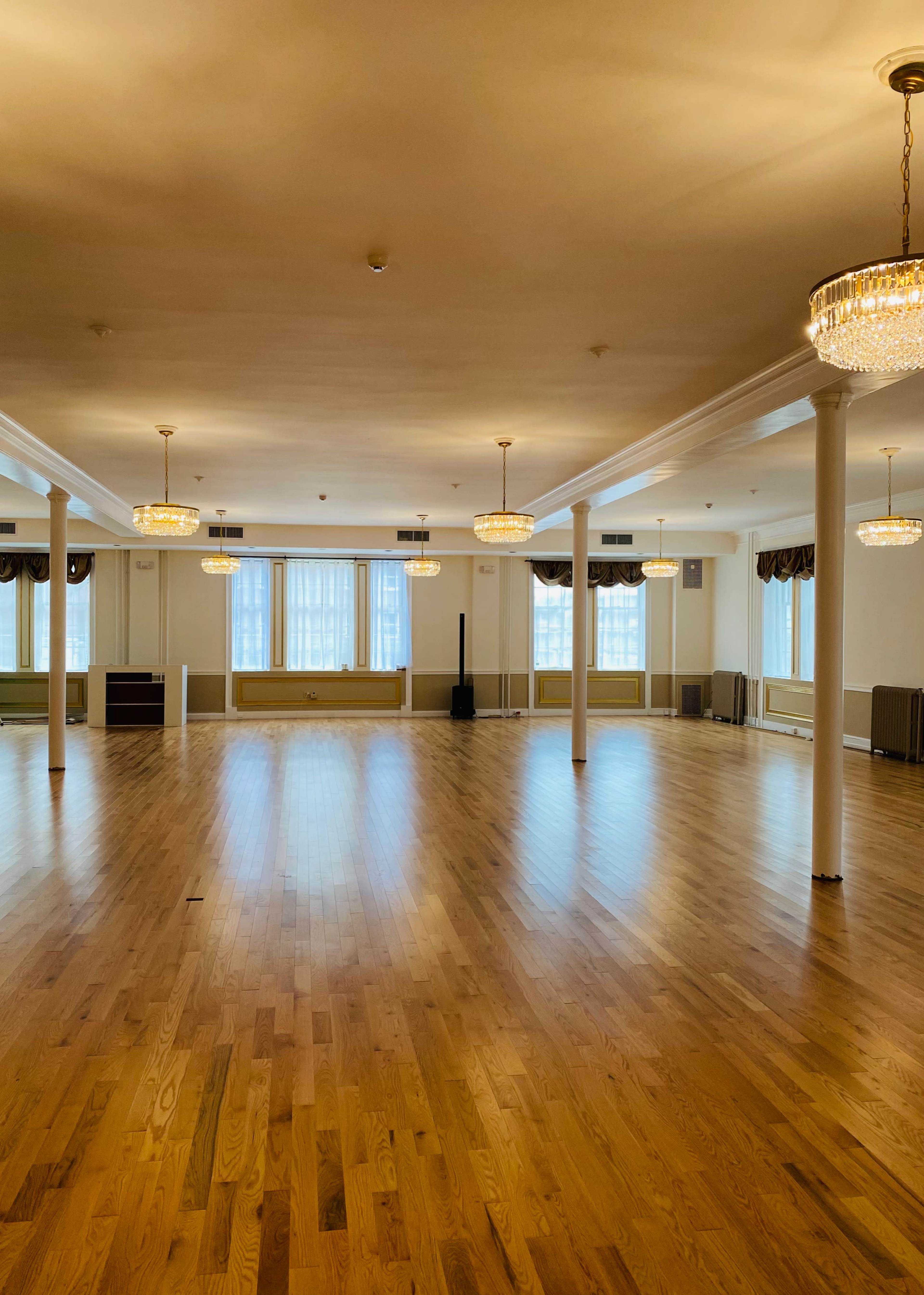 A spacious, well-lit hall features polished wooden floors, several chandeliers, and large windows along the walls.