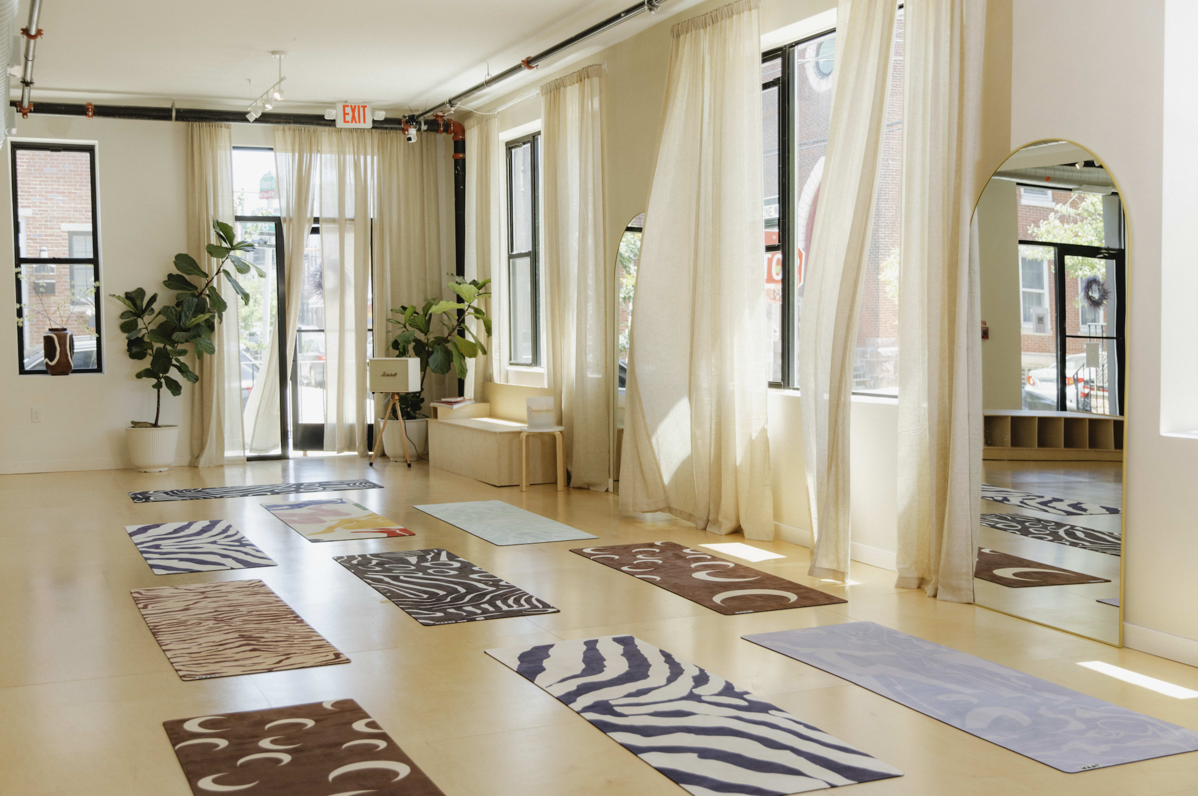 The image shows a bright yoga studio with several colorful mats arranged on the floor and large windows allowing natural light to enter.