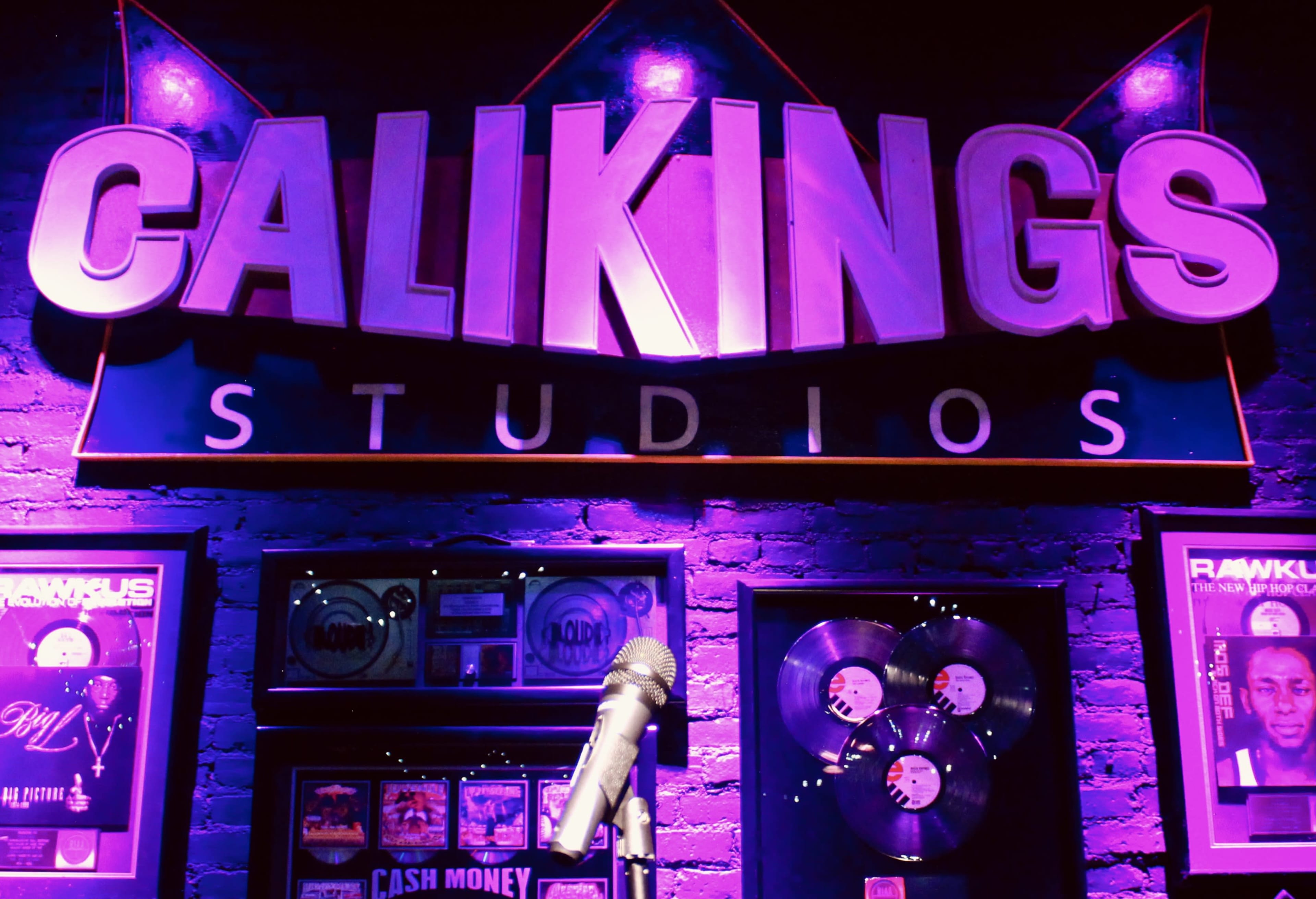 A wall displaying the "CaliKings Studios" sign, a microphone, and framed records under purple lighting.