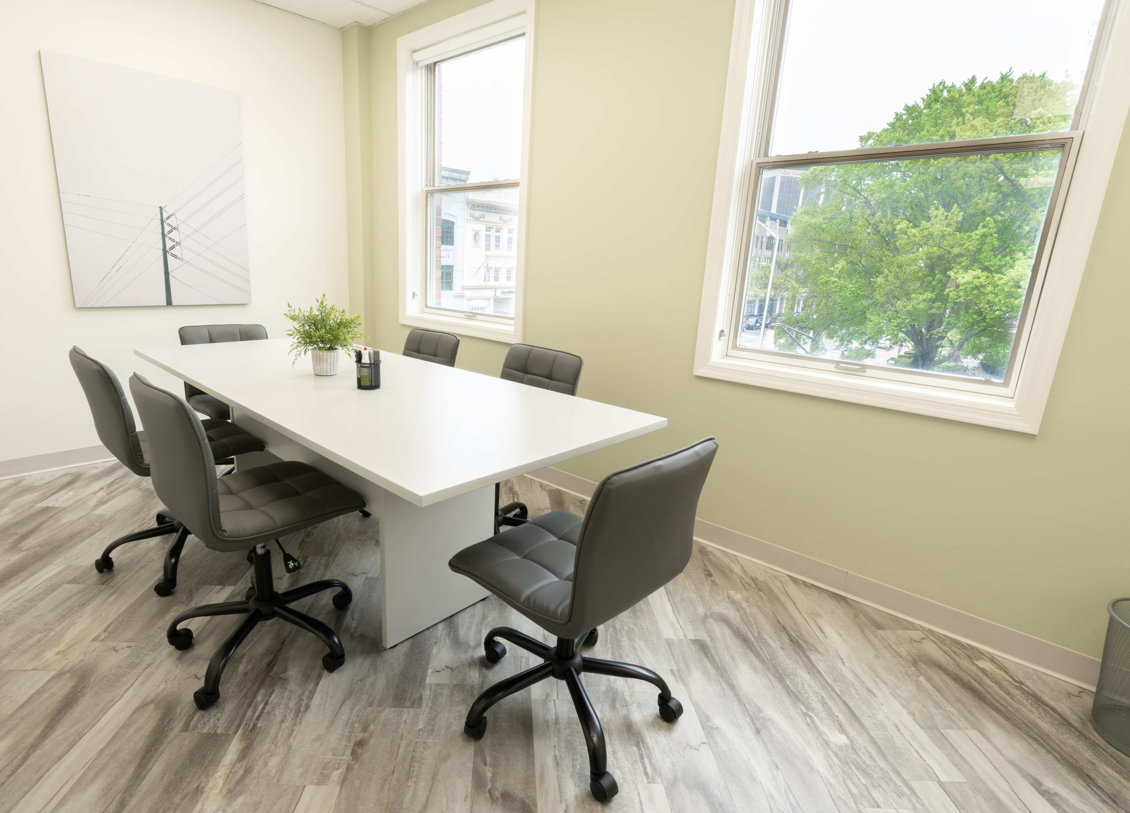 A modern conference room features a long white table surrounded by six gray office chairs, with large windows offering a view of greenery outside.
