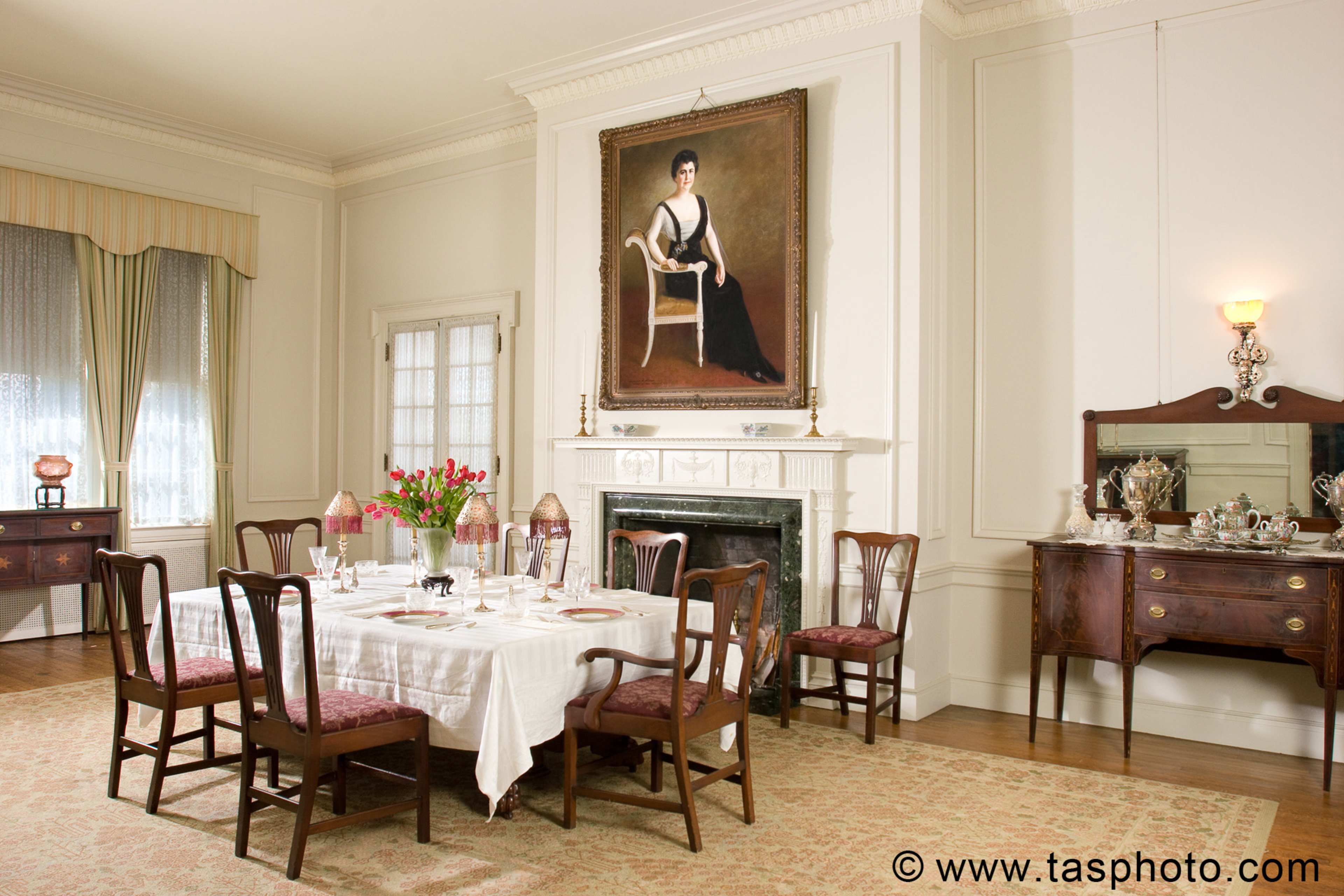 A dining room with a large table set for a meal, surrounded by eight wooden chairs, featuring a portrait of a woman on the wall and a sideboard displaying decorative items.