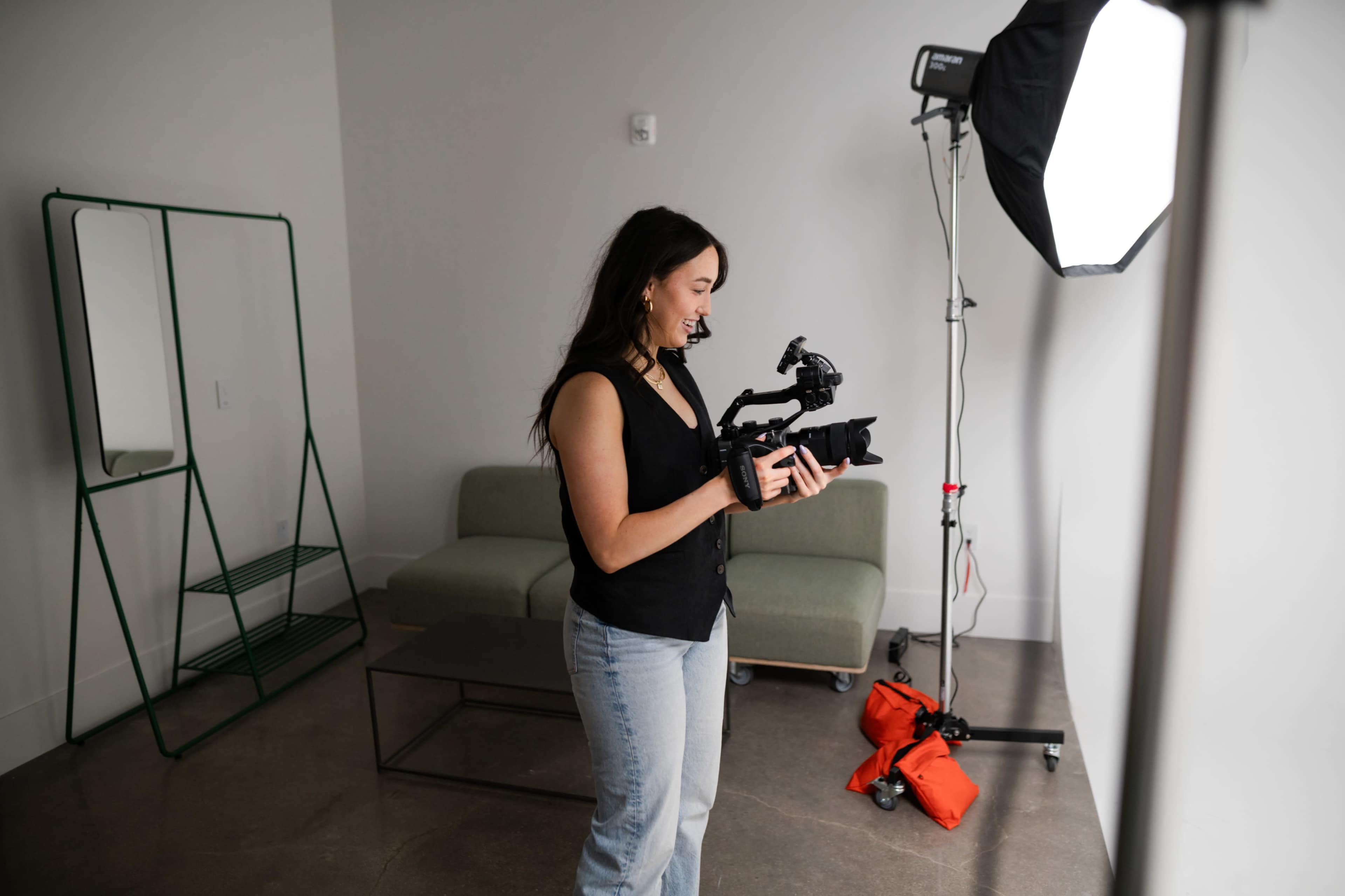 A woman stands holding a camera in a minimalist studio with a green couch and lighting equipment.