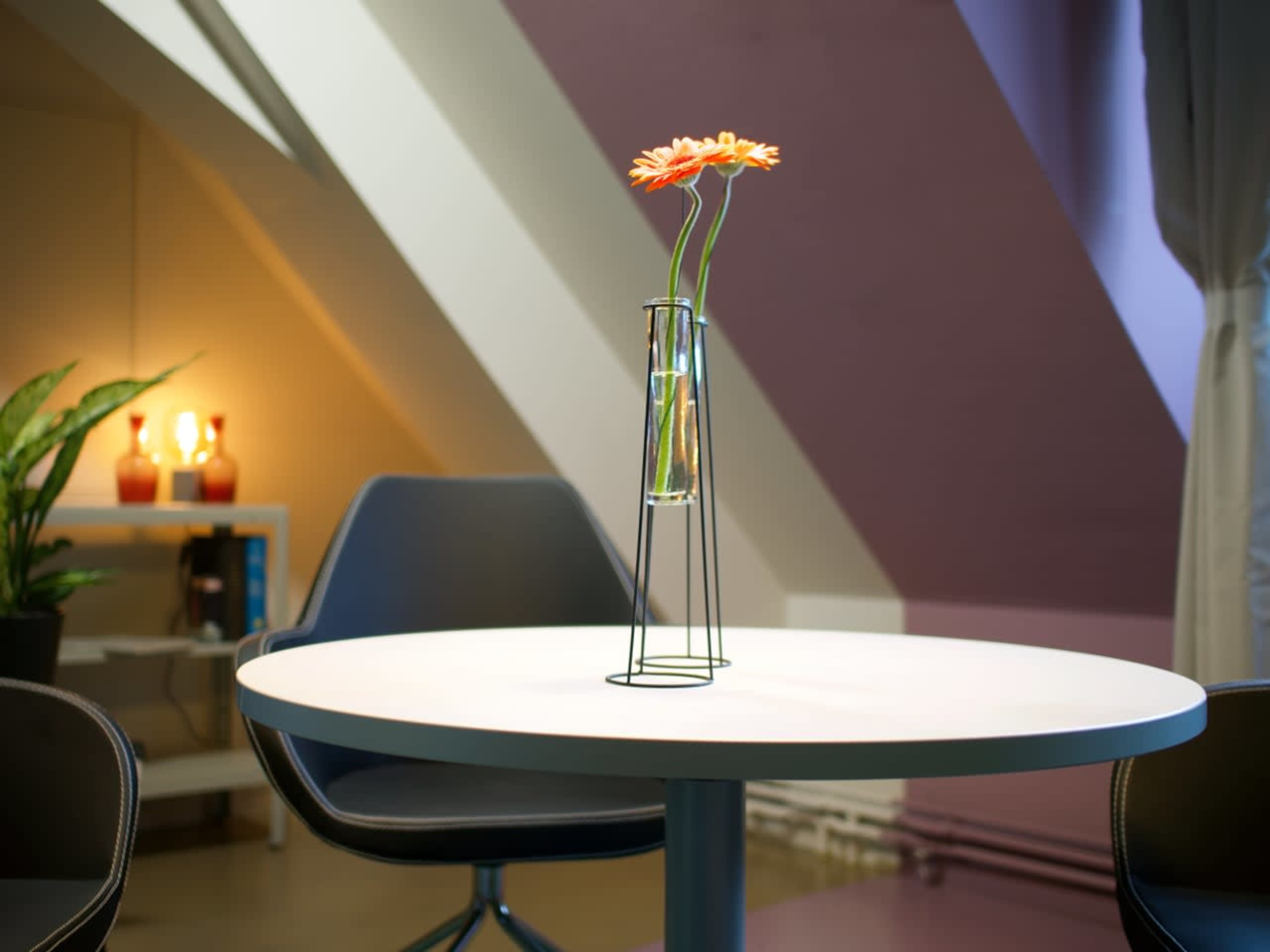 A minimalist round table with a single vase containing a flower is set in a room with sloped walls and soft lighting.
