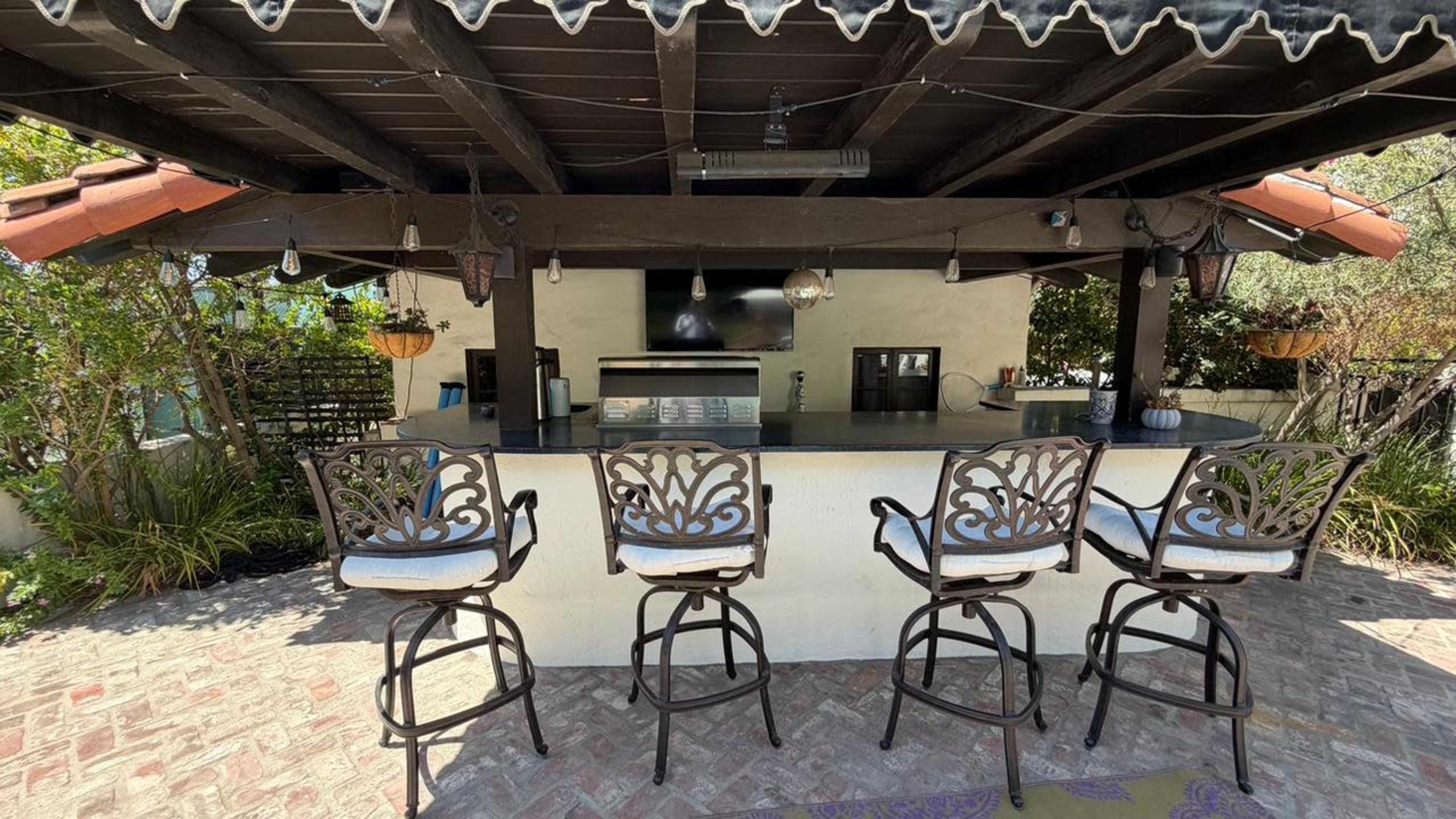 The image shows an outdoor bar area featuring a counter with four high-backed stools beneath a wooden roof structure.