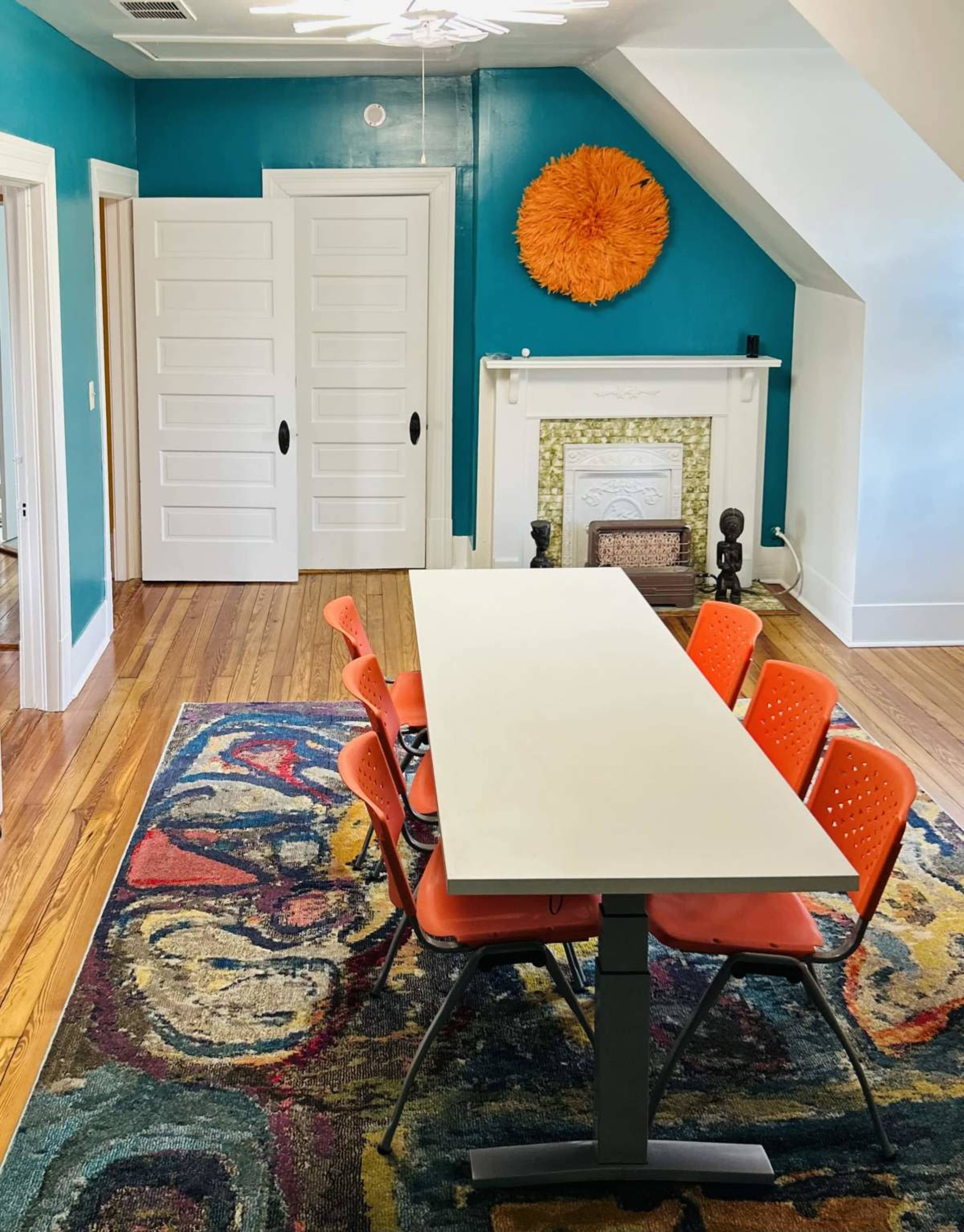 The image shows a dining area featuring a long white table surrounded by six orange chairs, set against a bright turquoise wall and a colorful area rug.