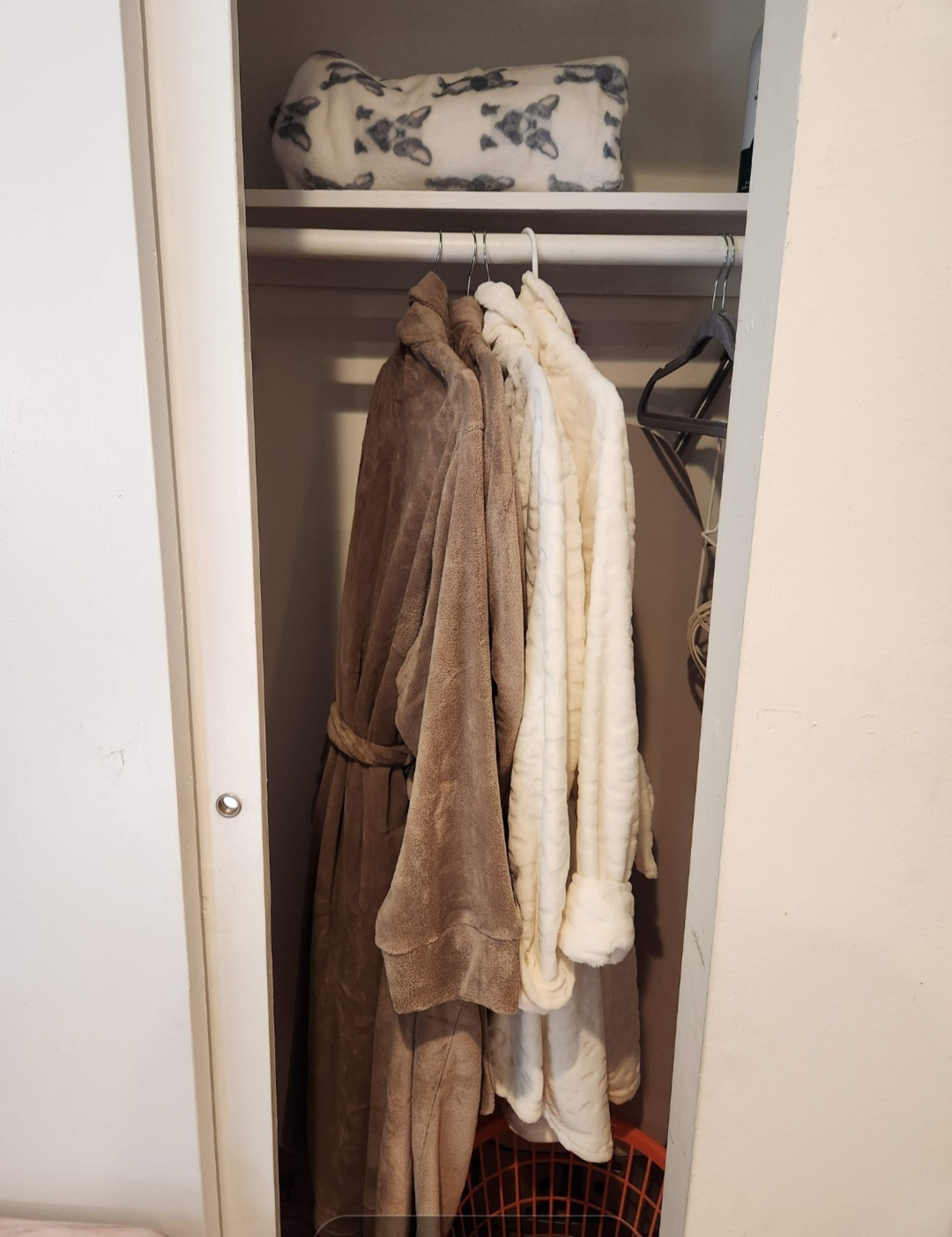 A closet contains three bathrobes hanging on a rod, with a blanket neatly folded on a shelf above.
