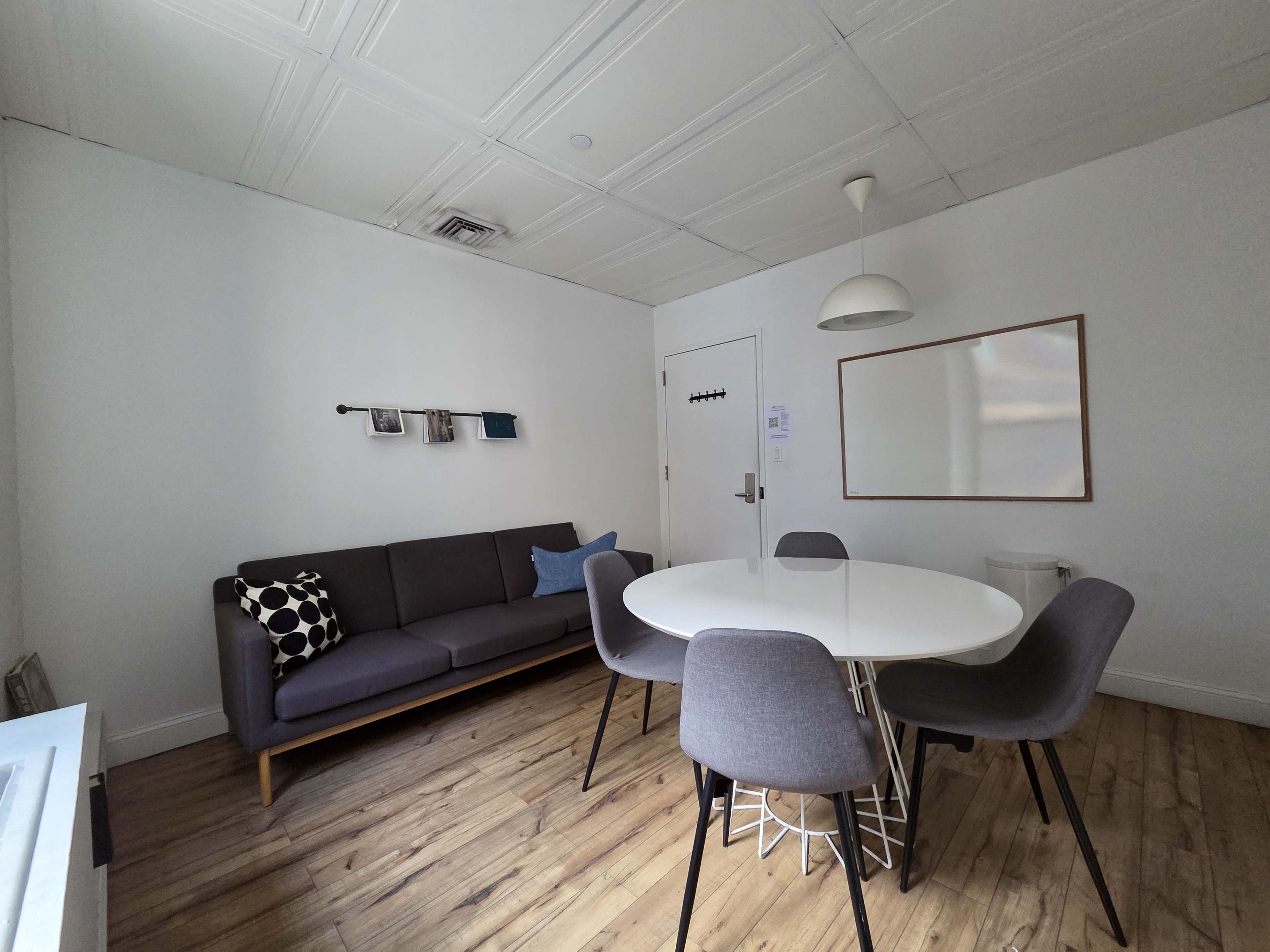The image shows a modern room featuring a gray sofa, a round white dining table surrounded by gray chairs, a wall mirror, and wooden flooring.