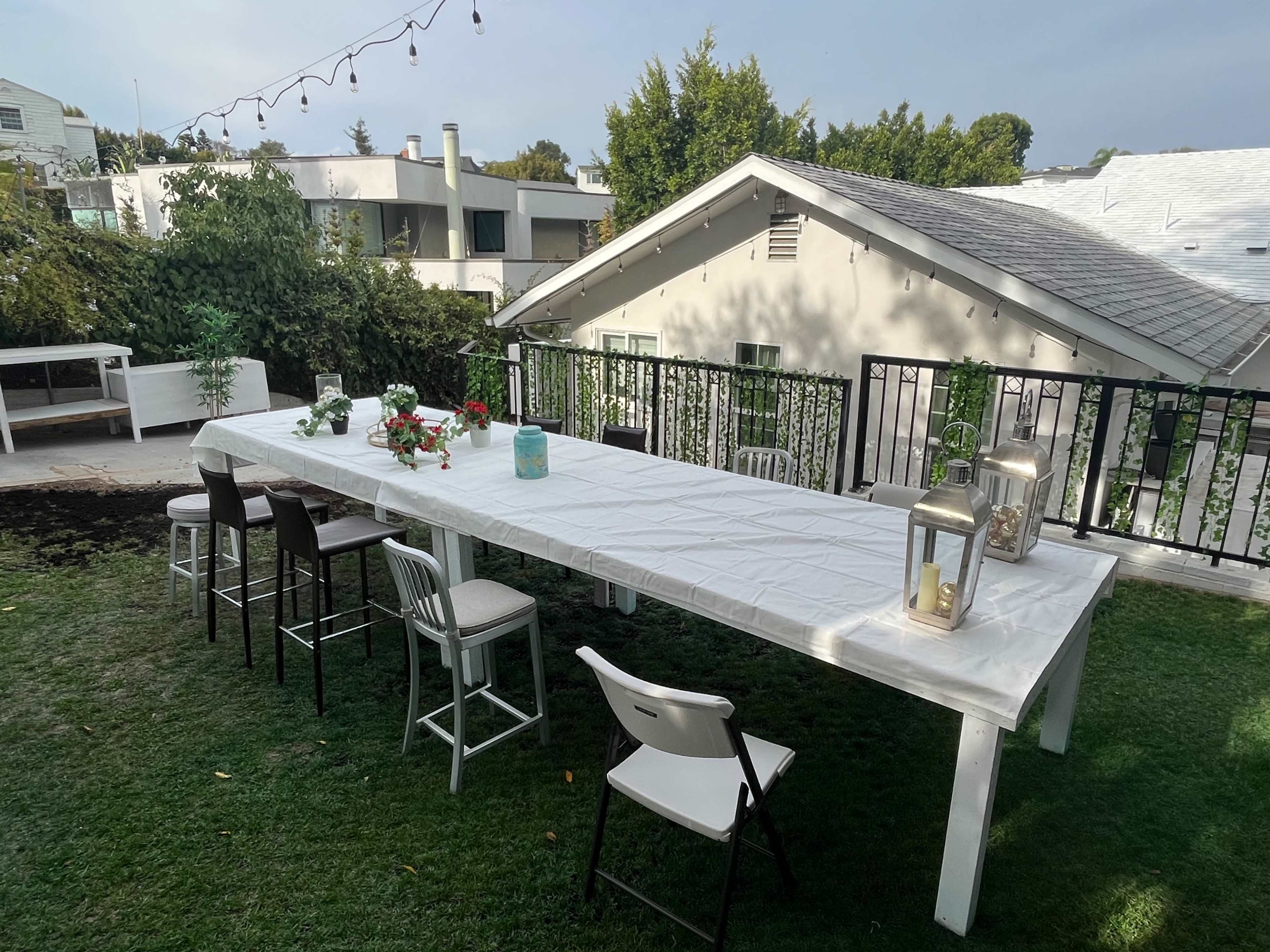 A long, white table is set outdoors on grass, surrounded by various chairs, with decorative items including lanterns and flowers.