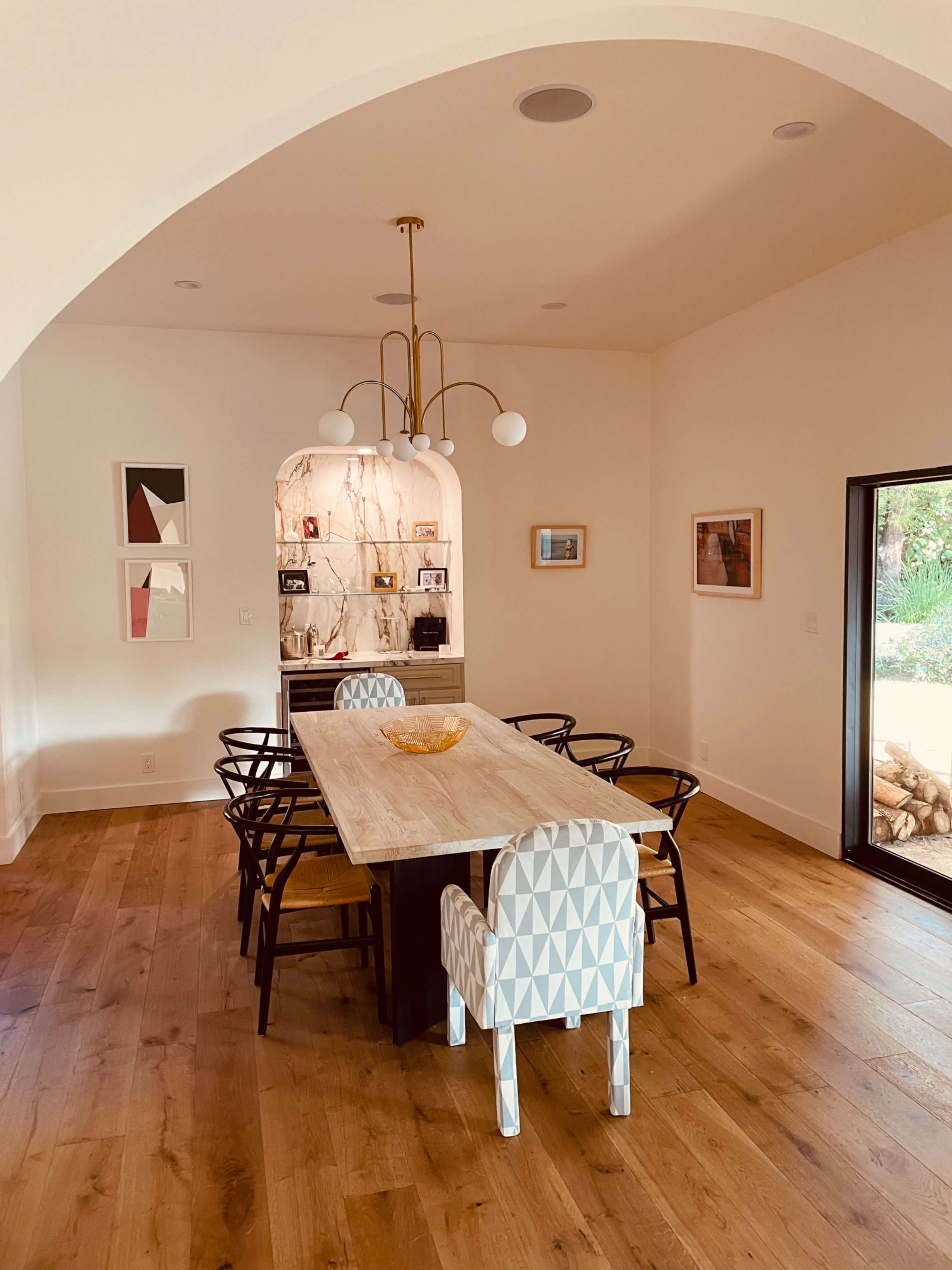 A dining room features a long wooden table surrounded by six chairs, with a light fixture hanging above and a window showcasing a view outside.