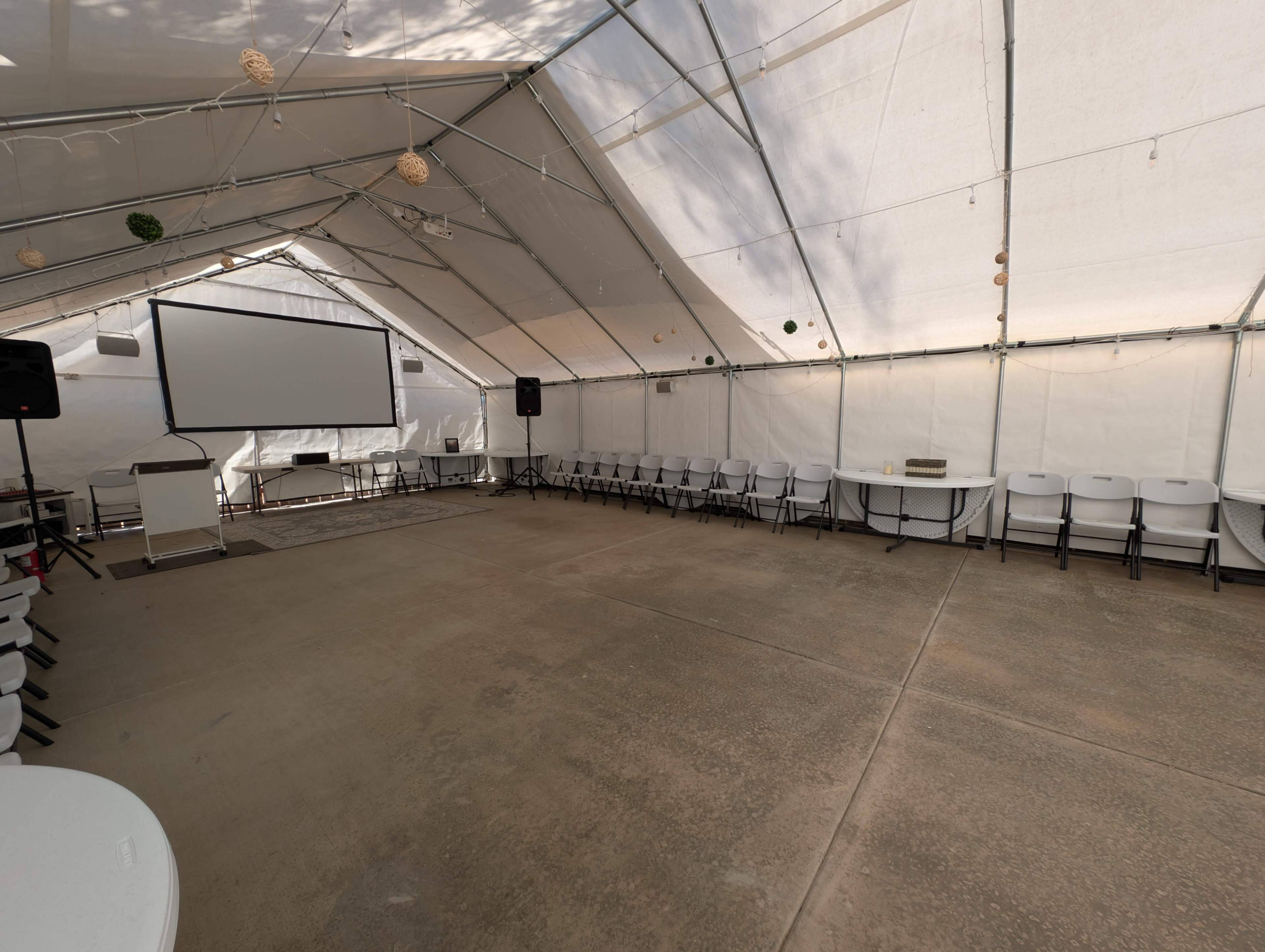 The image shows an indoor tent setup with rows of folding chairs, a projector screen, and a speaker system.