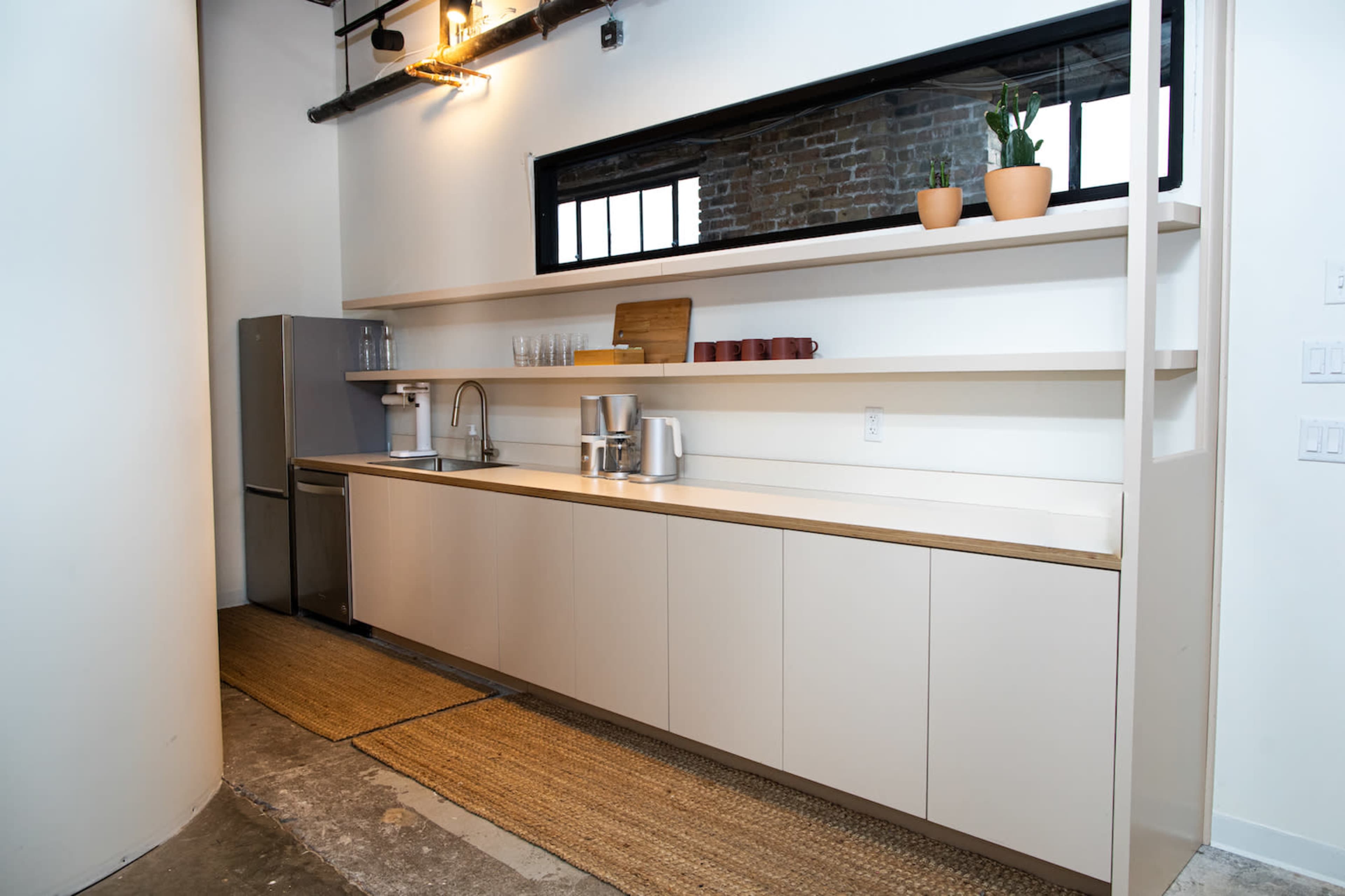 The image shows a minimalist kitchen with a long white countertop, shelves displaying dishes and a plant, and a stainless steel refrigerator.